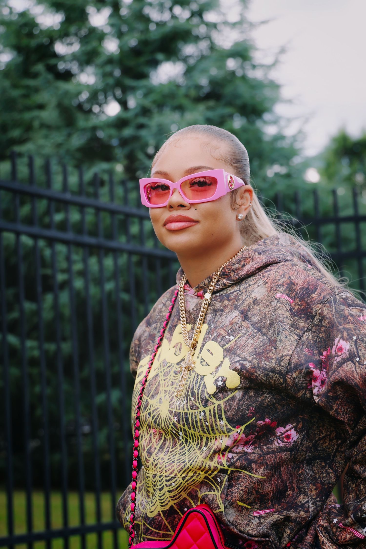 Stylish woman with shaved head wearing pink cat-eye sunglasses, floral hoodie, and gold chain necklace outdoors.