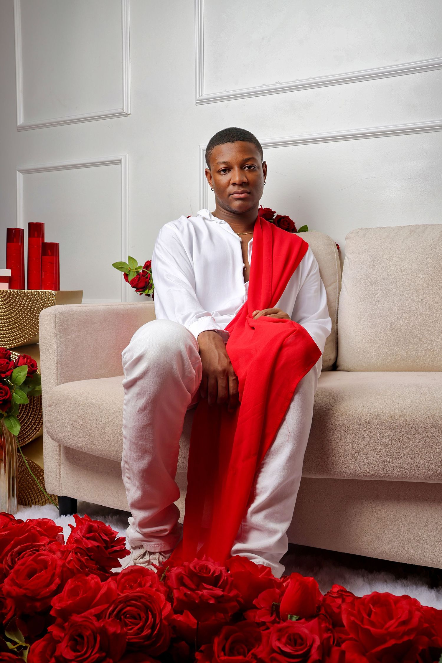 Young man in white outfit with red sash sits on beige sofa surrounded by red roses in elegant white room.