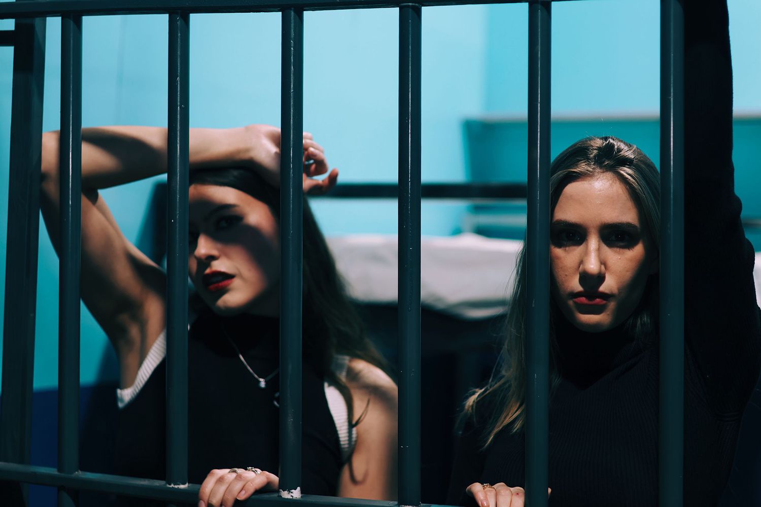 Two women with dark hair stand behind jail cell bars in a dramatic, moody blue-lit prison scene.