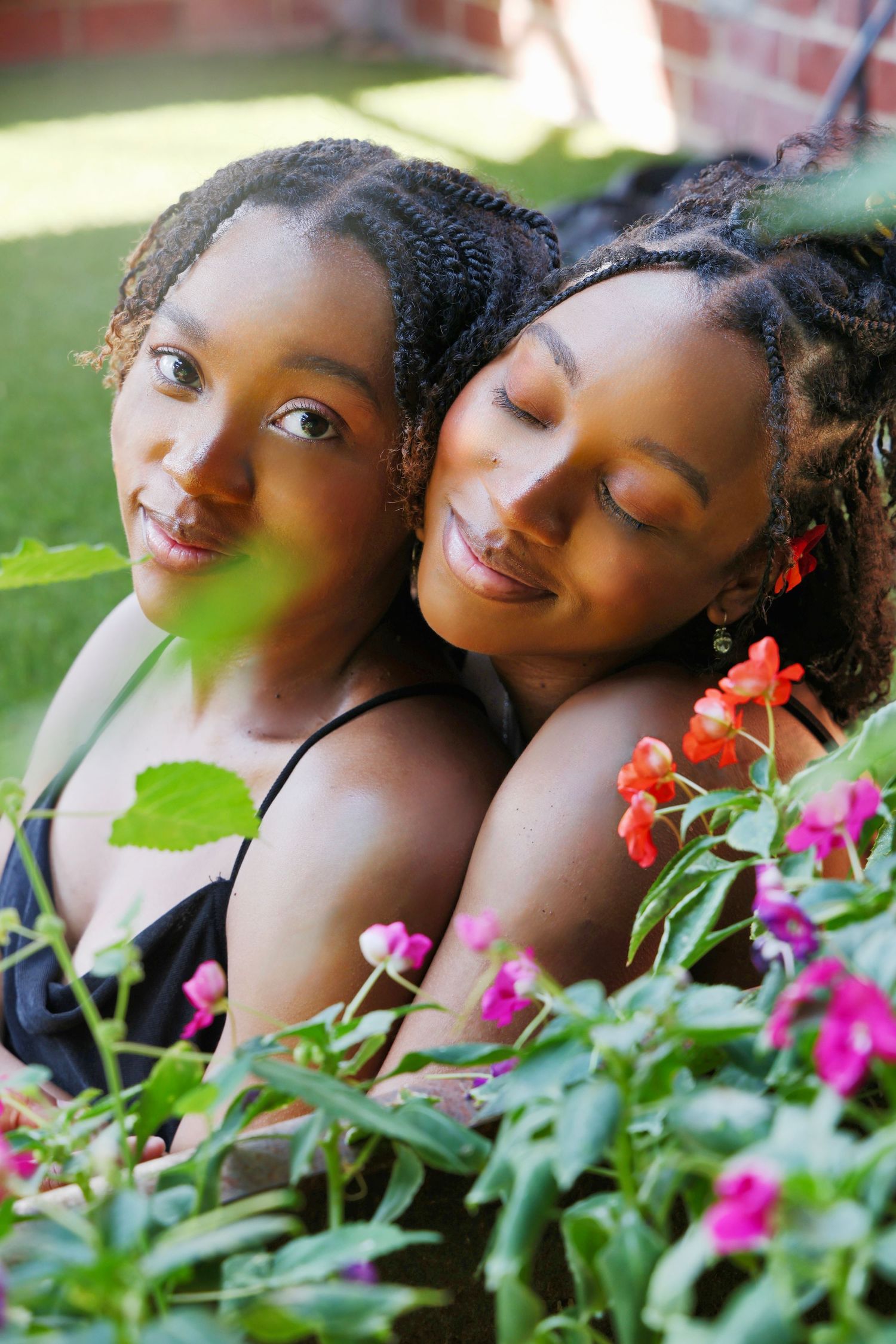 Two young Black women smiling together among colorful garden flowers, candid outdoor portrait.