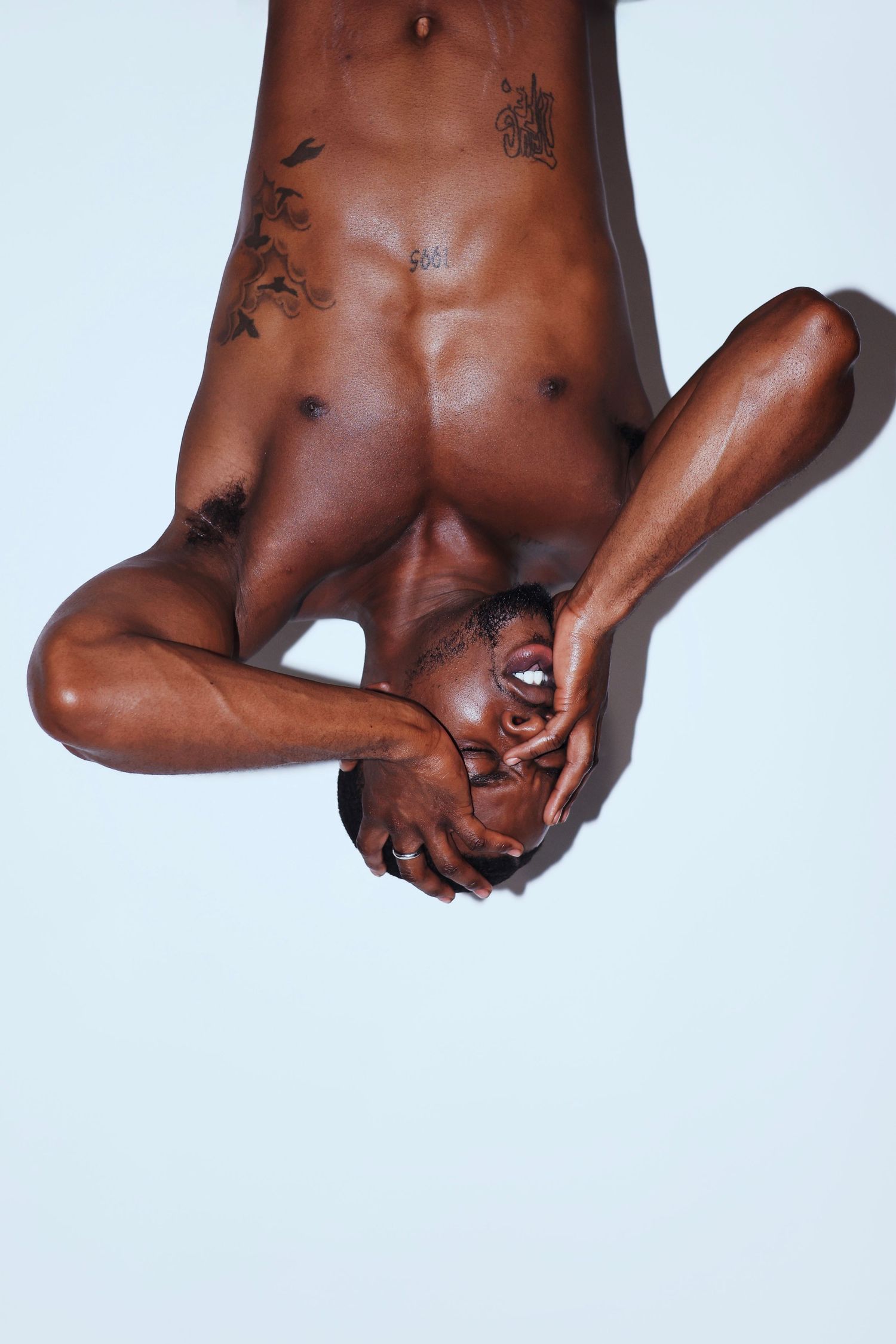 Shirtless tattooed Black man photographed upside down against a light blue background in an artistic pose.