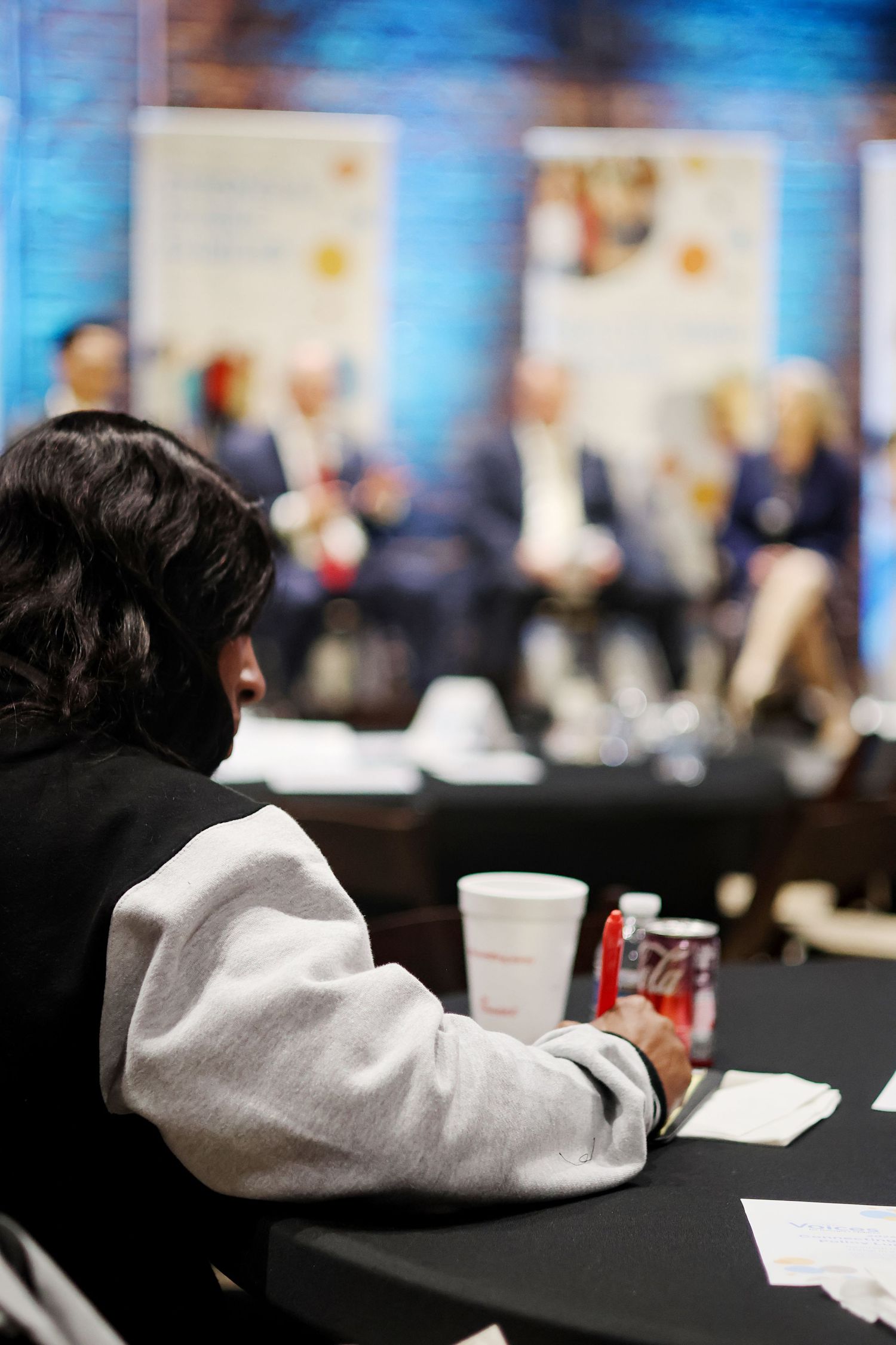 Audience member at a conference panel event, sitting at a table with drinks and papers, panel speakers blurred in background.