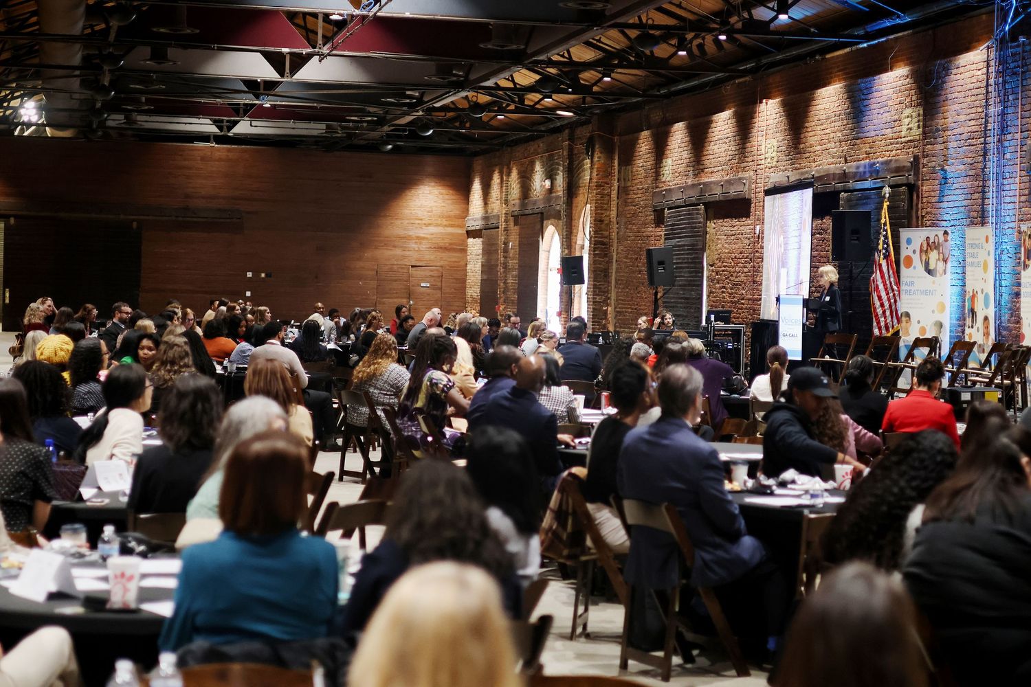 Large conference event in a brick-walled venue with attendees seated at tables listening to a speaker on stage.