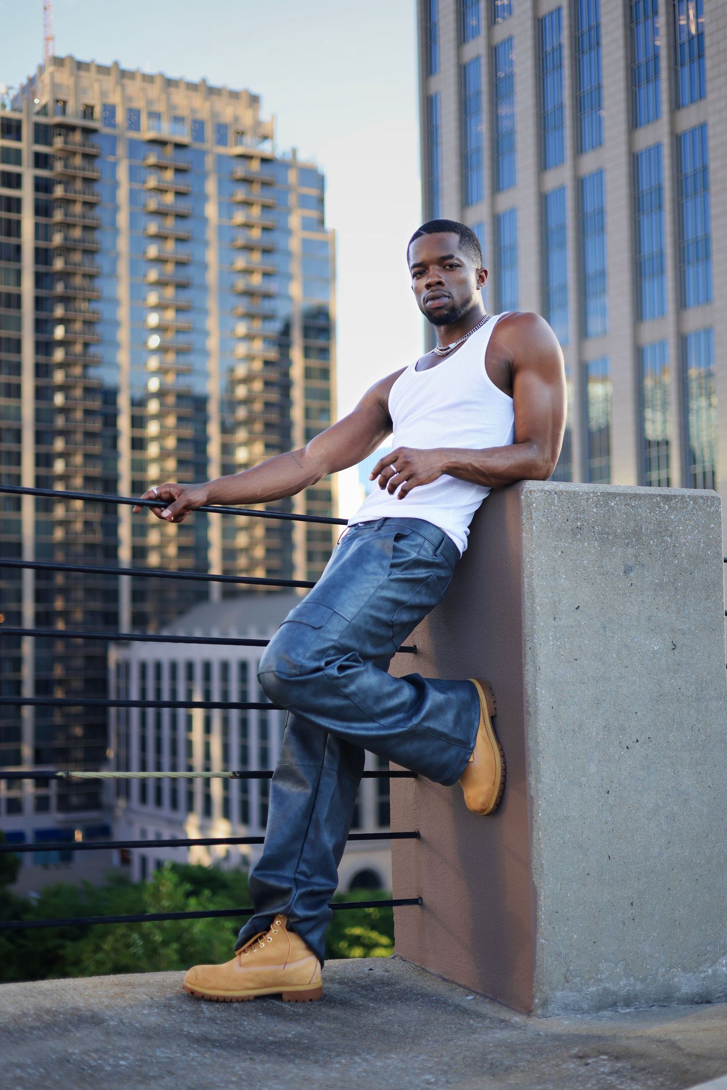Athletic man in white tank top and jeans posing against rooftop wall with urban skyline.
