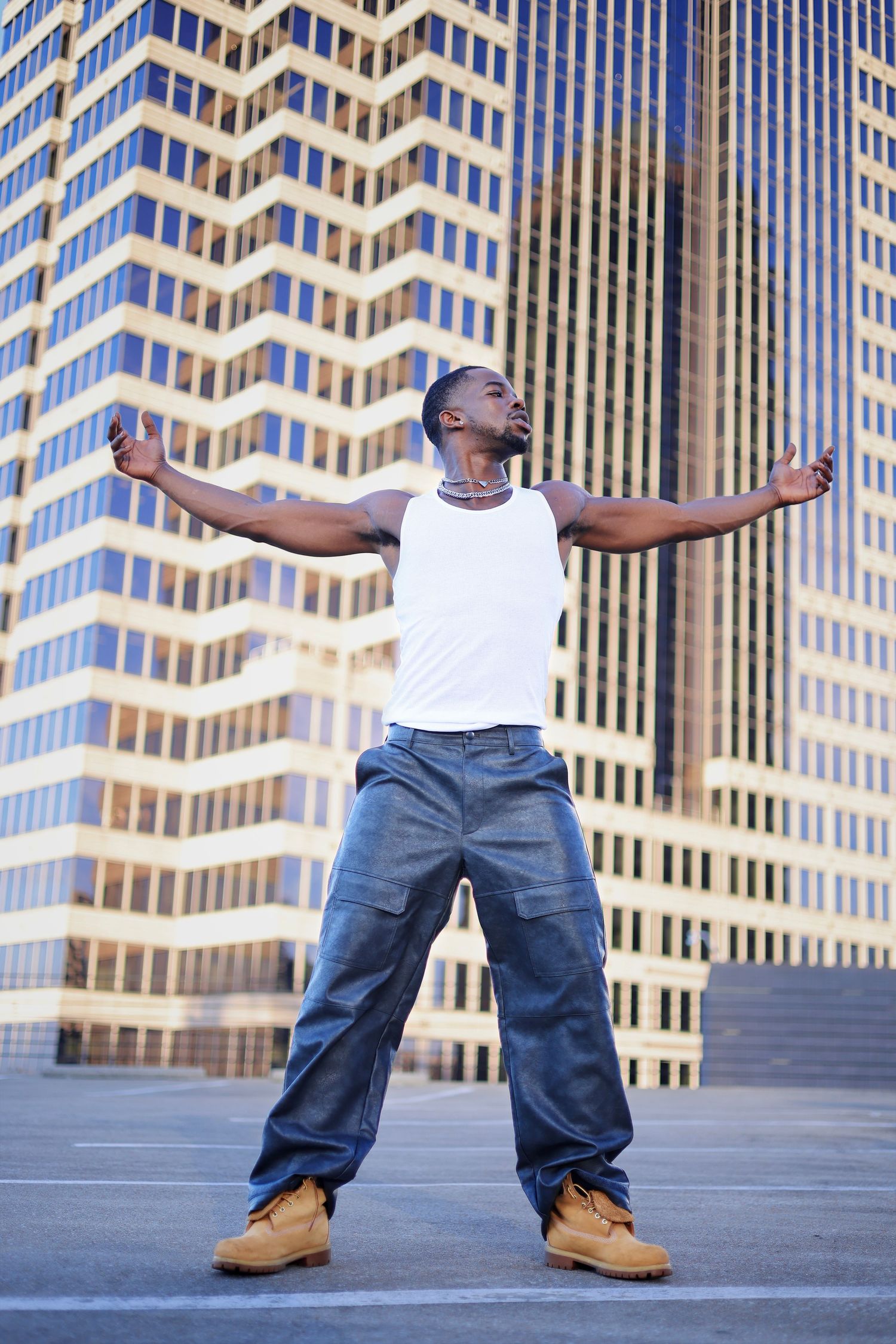 Confident Black man in white tank top spreading arms wide in front of golden skyscraper.