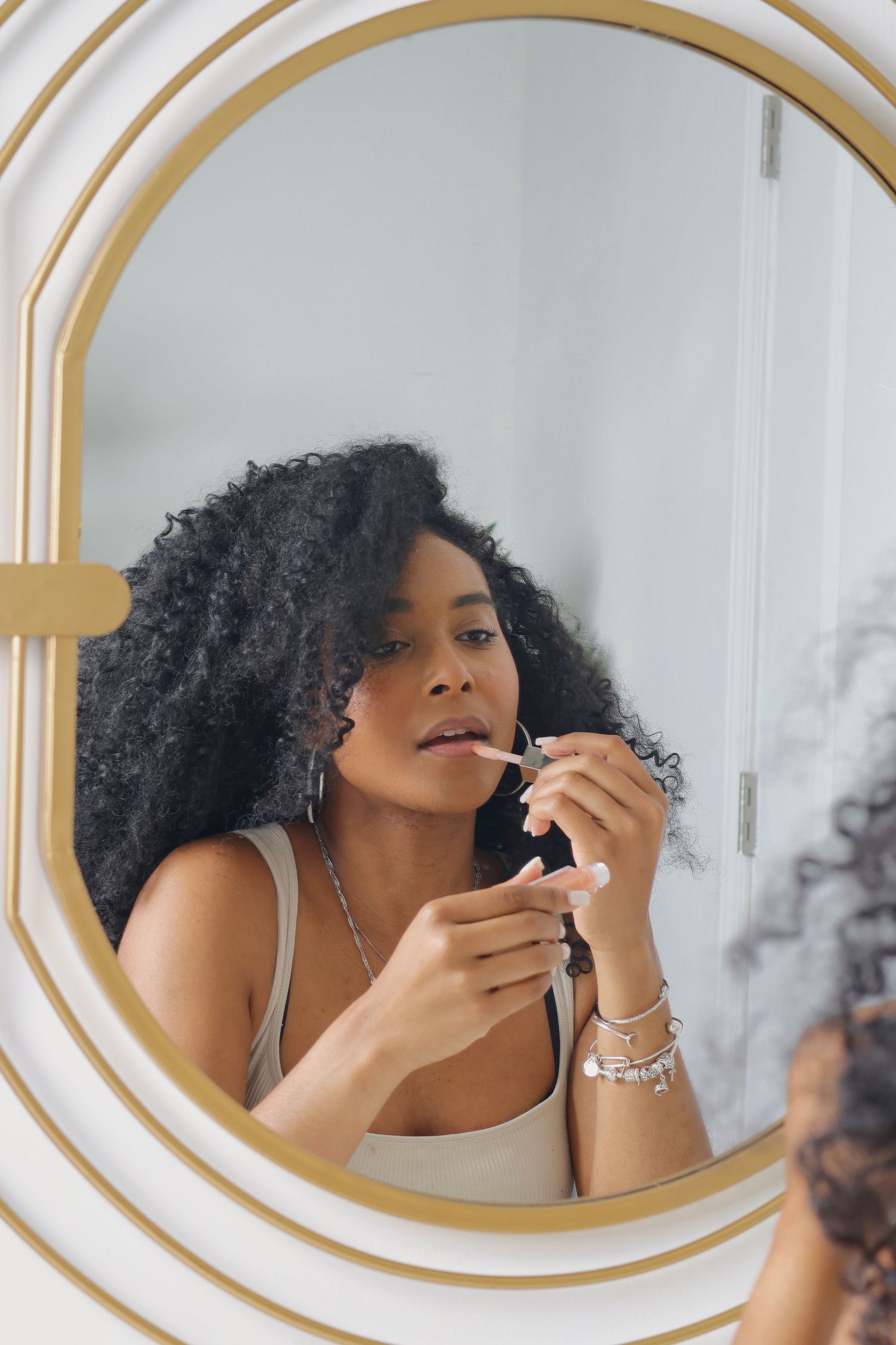 Young Black woman applying lip gloss while looking at her reflection in a round gold-framed mirror.