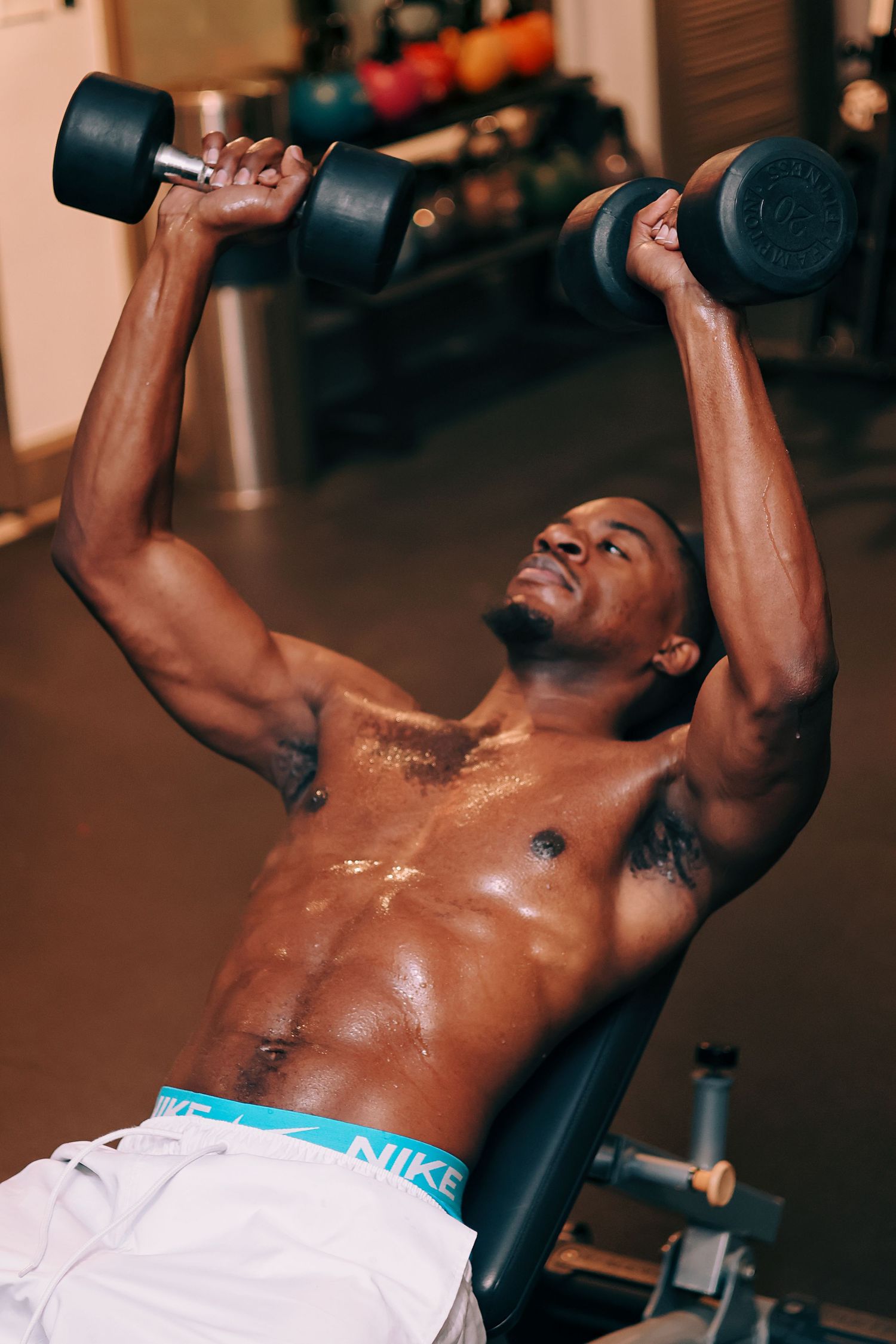 Muscular shirtless Black man performing dumbbell chest press on incline bench, wearing white shorts and Nike underwear.