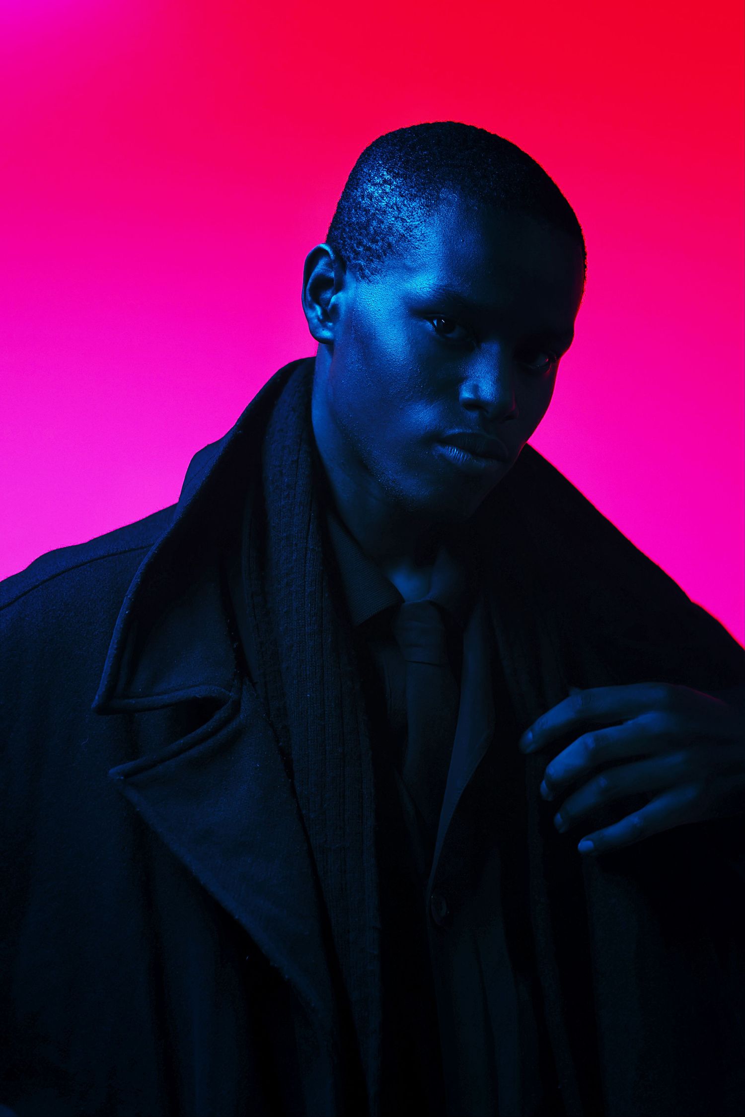 A Black man in a dark trench coat poses against a vibrant pink background with dramatic blue lighting.