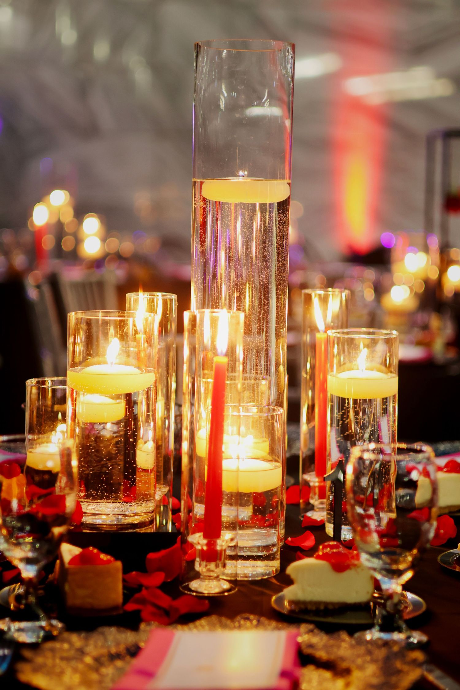 Elegant wedding reception centerpiece with floating candles in tall glass cylinders and red rose petals on a decorated table.