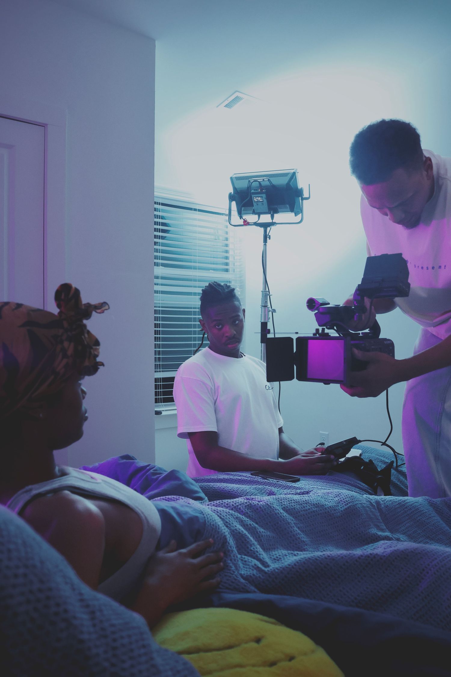 Behind-the-scenes video shoot with crew filming a person lying in bed under purple studio lighting.