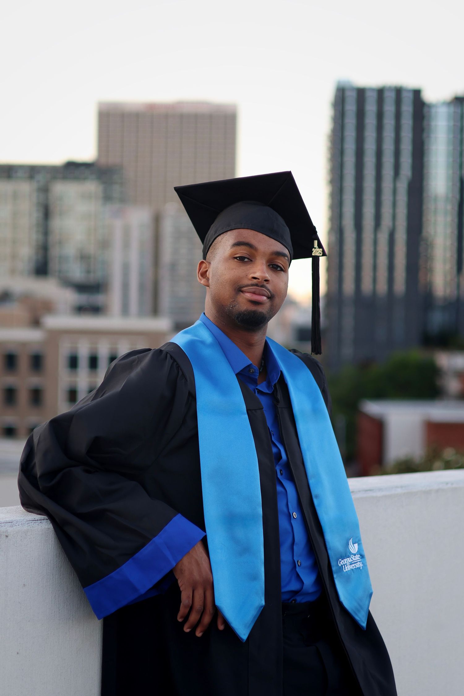 Young man in black graduation gown and blue stole poses on rooftop with city skyline backdrop.
