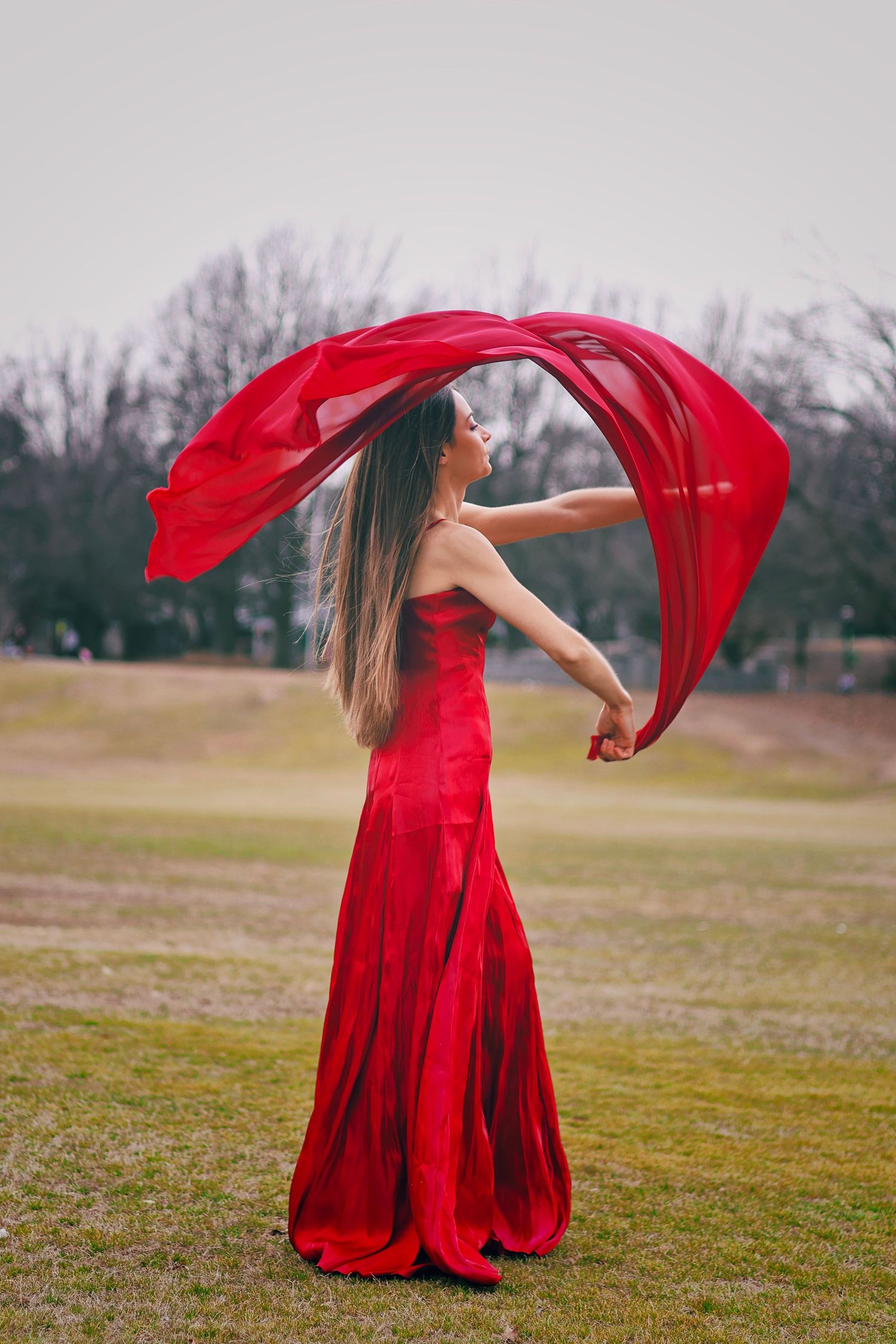 Woman in red strapless gown with flowing fabric billowing in wind outdoors in park.