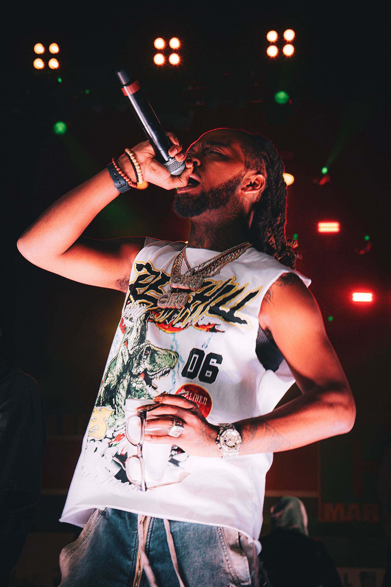 Male rapper performing on stage holding microphone wearing white graphic tank top and gold chains under colorful stage lights.
