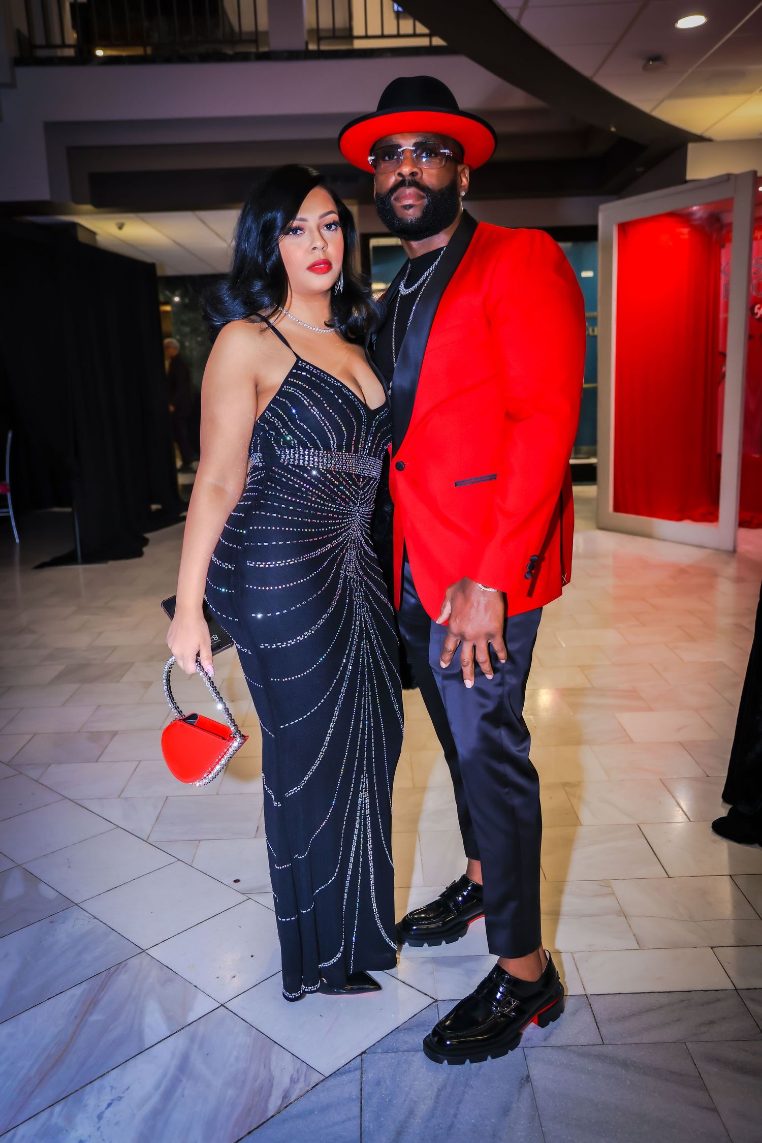 Elegant couple posing at formal event, woman in black beaded gown, man in red blazer and hat.