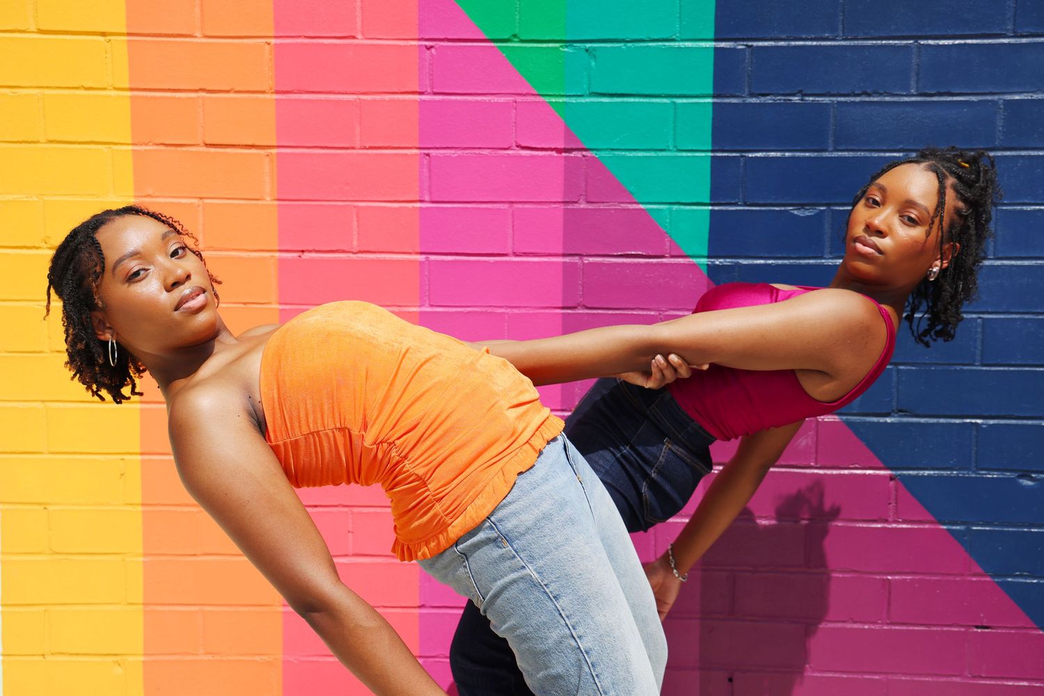 Two young Black women pose back-to-back against a vibrant rainbow-painted brick wall, wearing orange and pink tops.