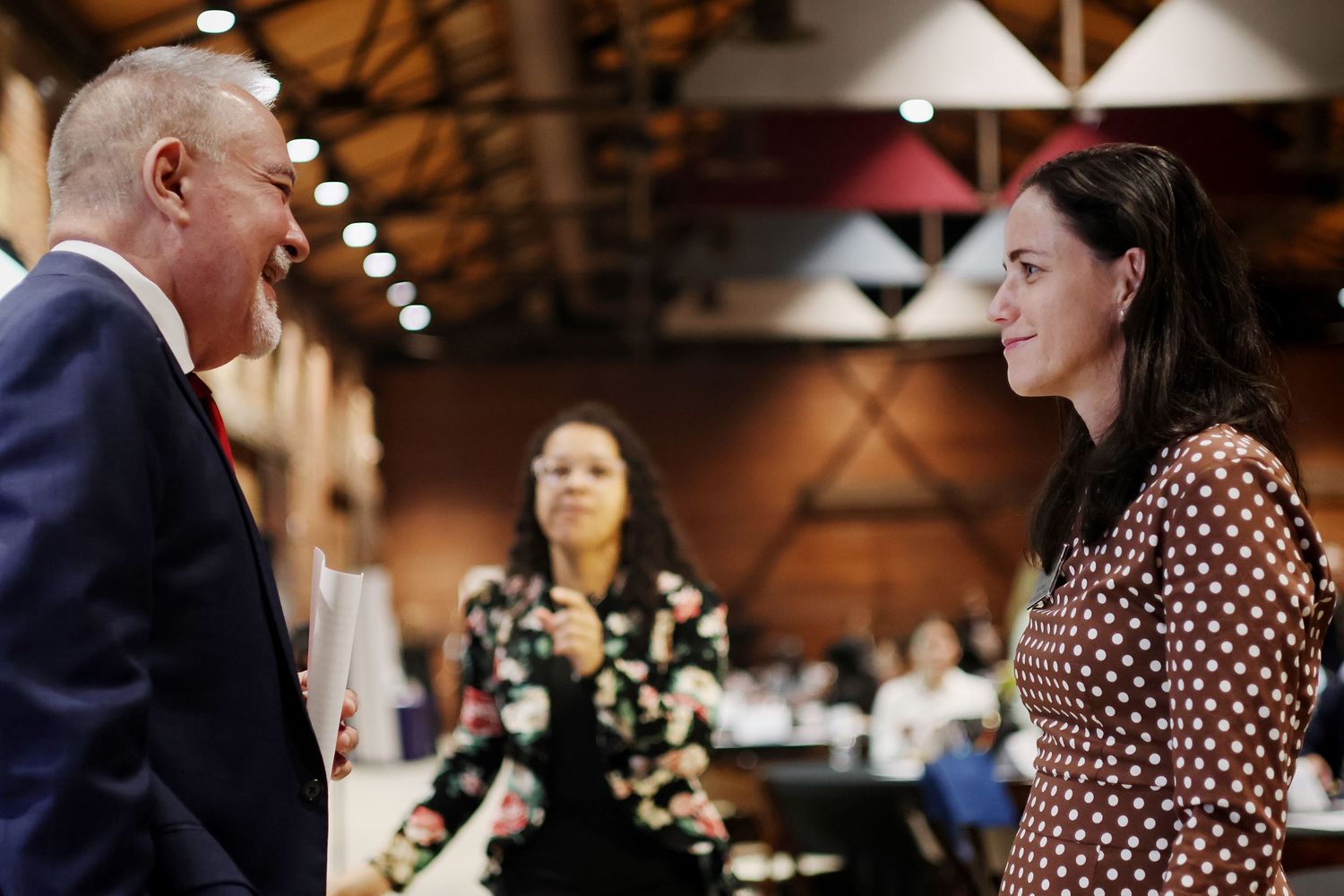 Two professionals engaged in conversation at a networking event, with attendees visible in the background.