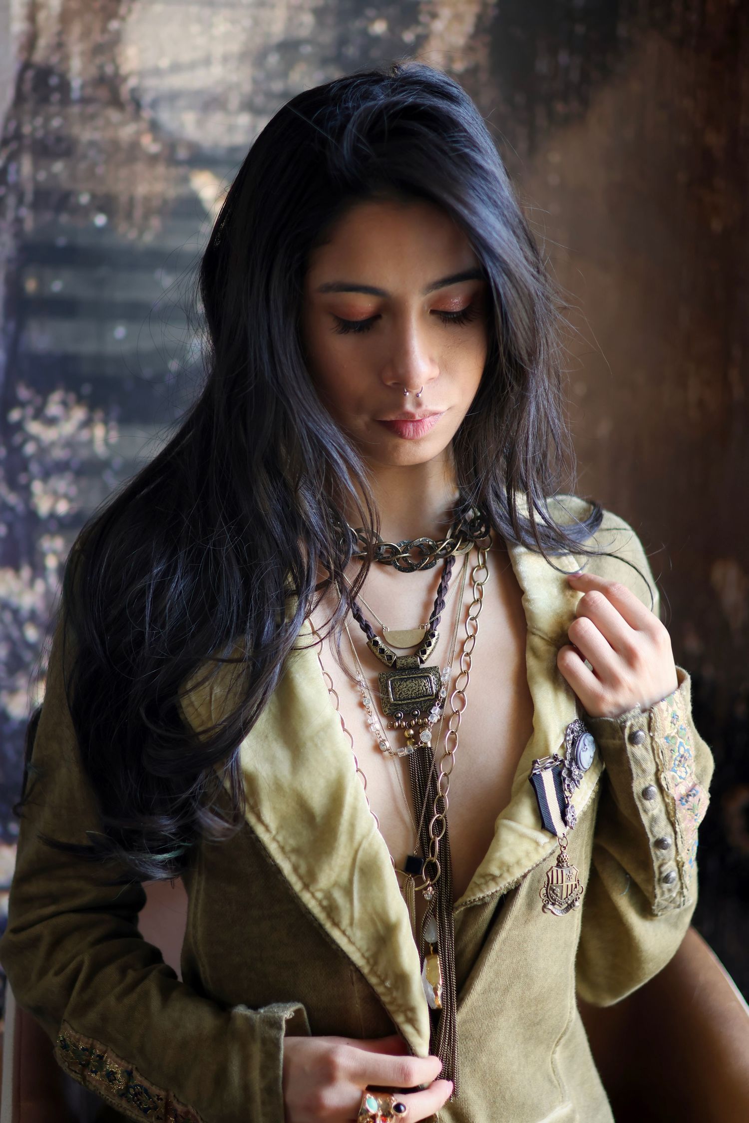 Woman with long dark hair in olive jacket wearing layered chain necklaces in moody editorial.