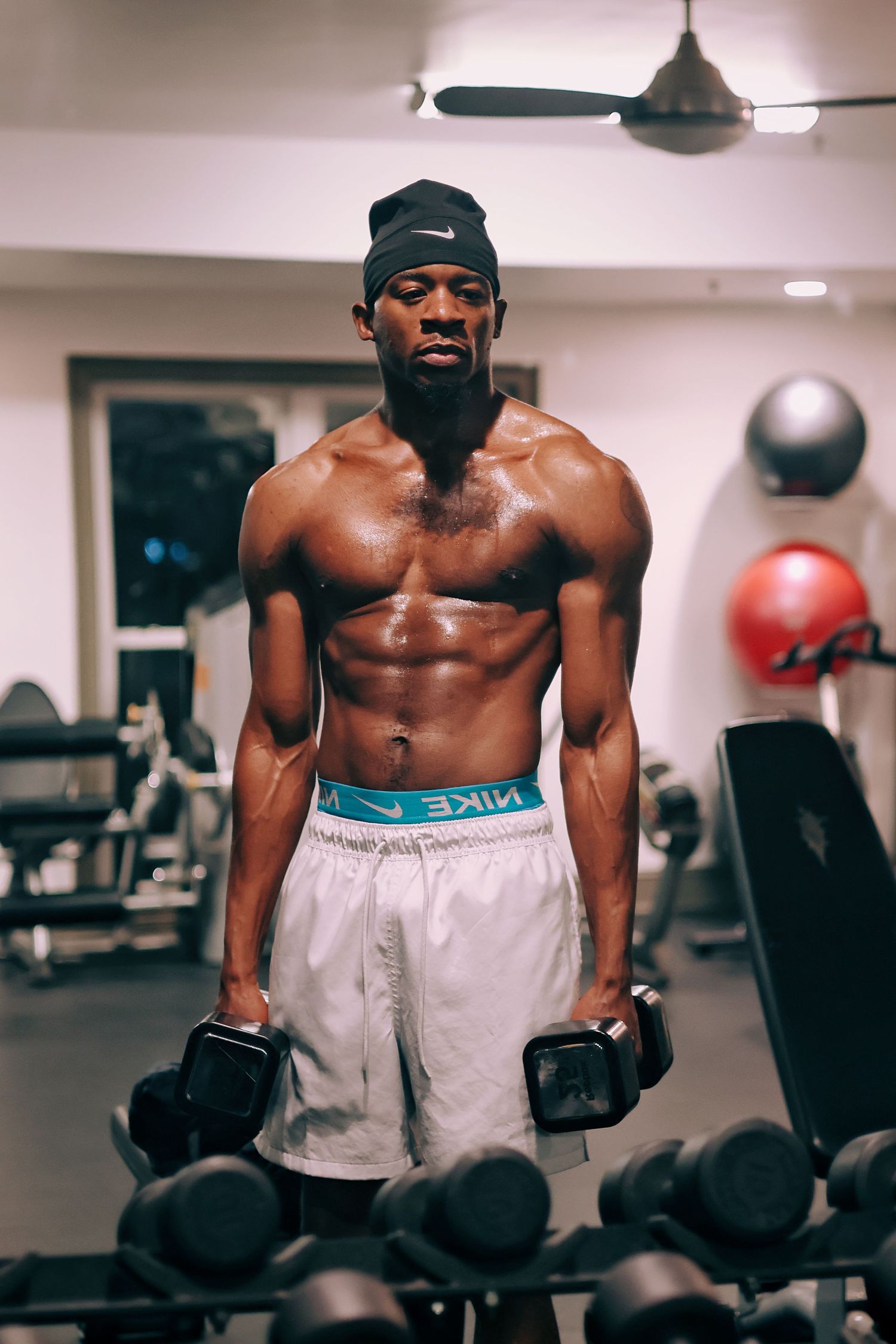 Muscular shirtless Black man holding dumbbells in gym wearing Nike shorts and black beanie.