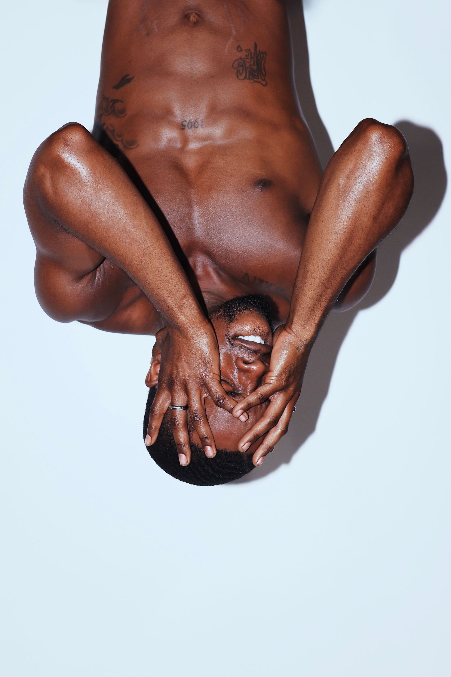 Shirtless athletic Black man with tattoos lying on light blue background, viewed from above with hands covering face.
