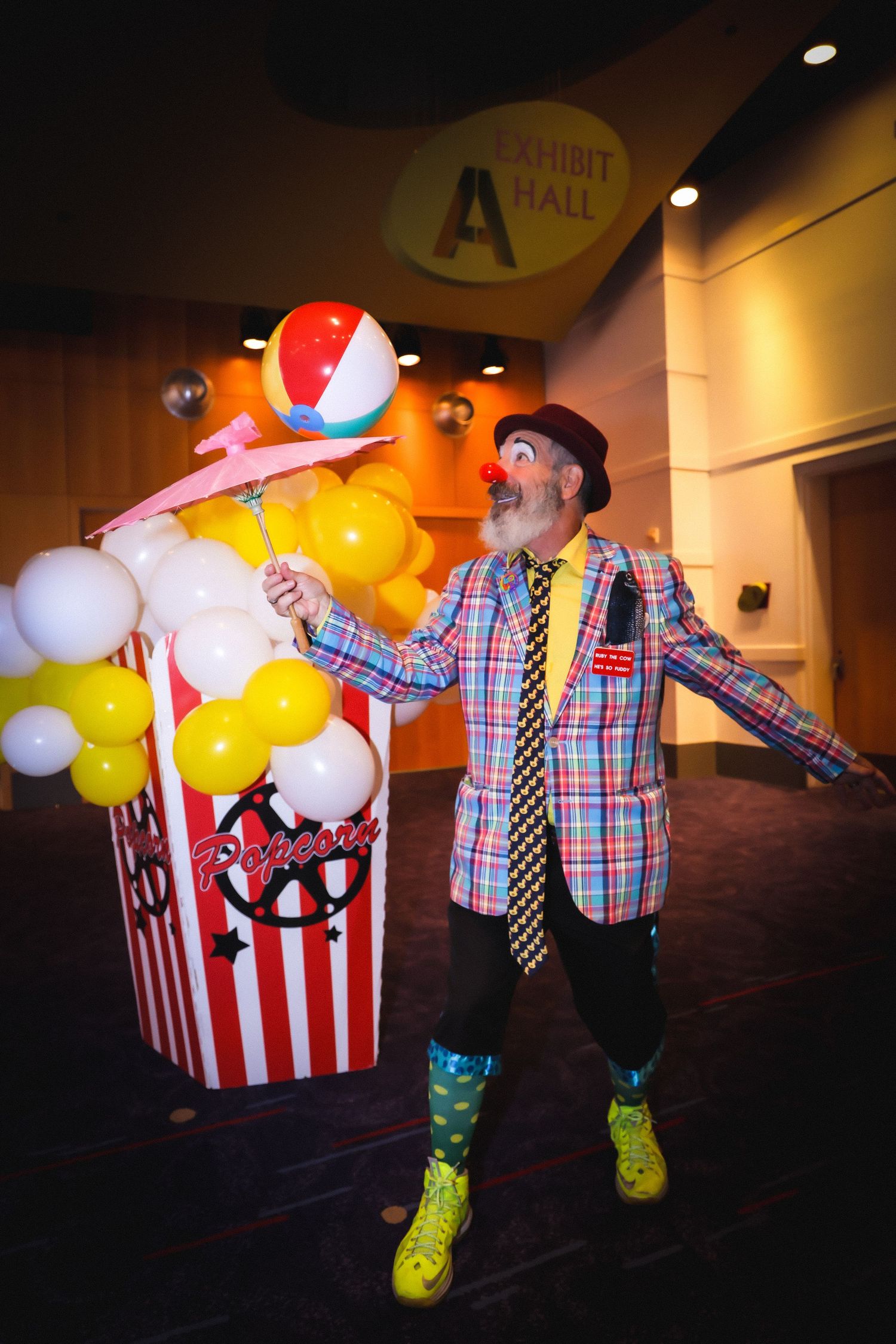 A colorful clown with red nose and plaid jacket poses with balloon popcorn display near Exhibit Hall A entrance.