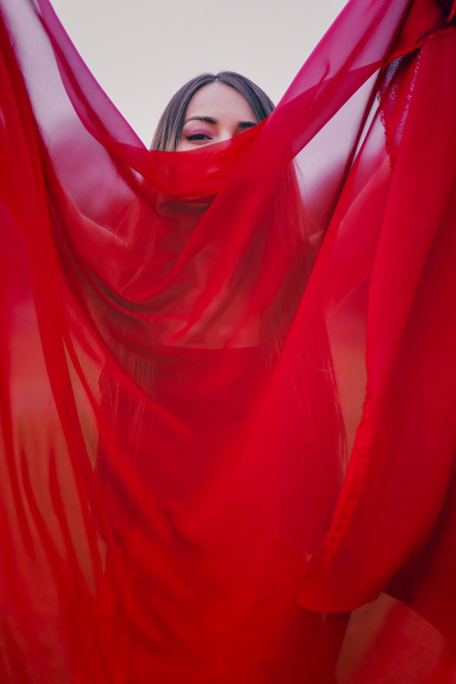 Woman holding flowing red sheer fabric dramatically around her face against a neutral background.