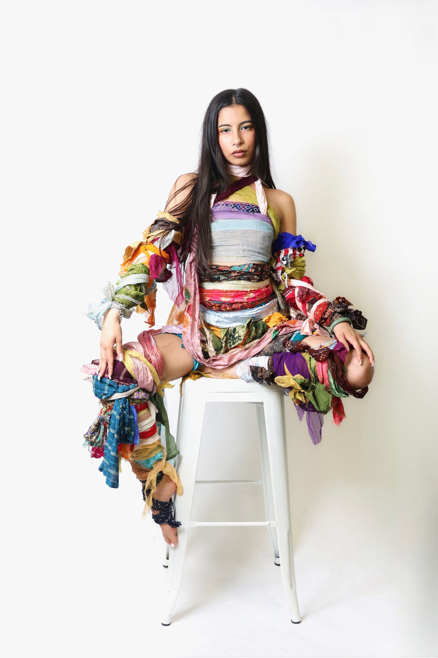 Woman sitting on white stool wearing colorful patchwork outfit with flowing fabric pieces in studio setting.