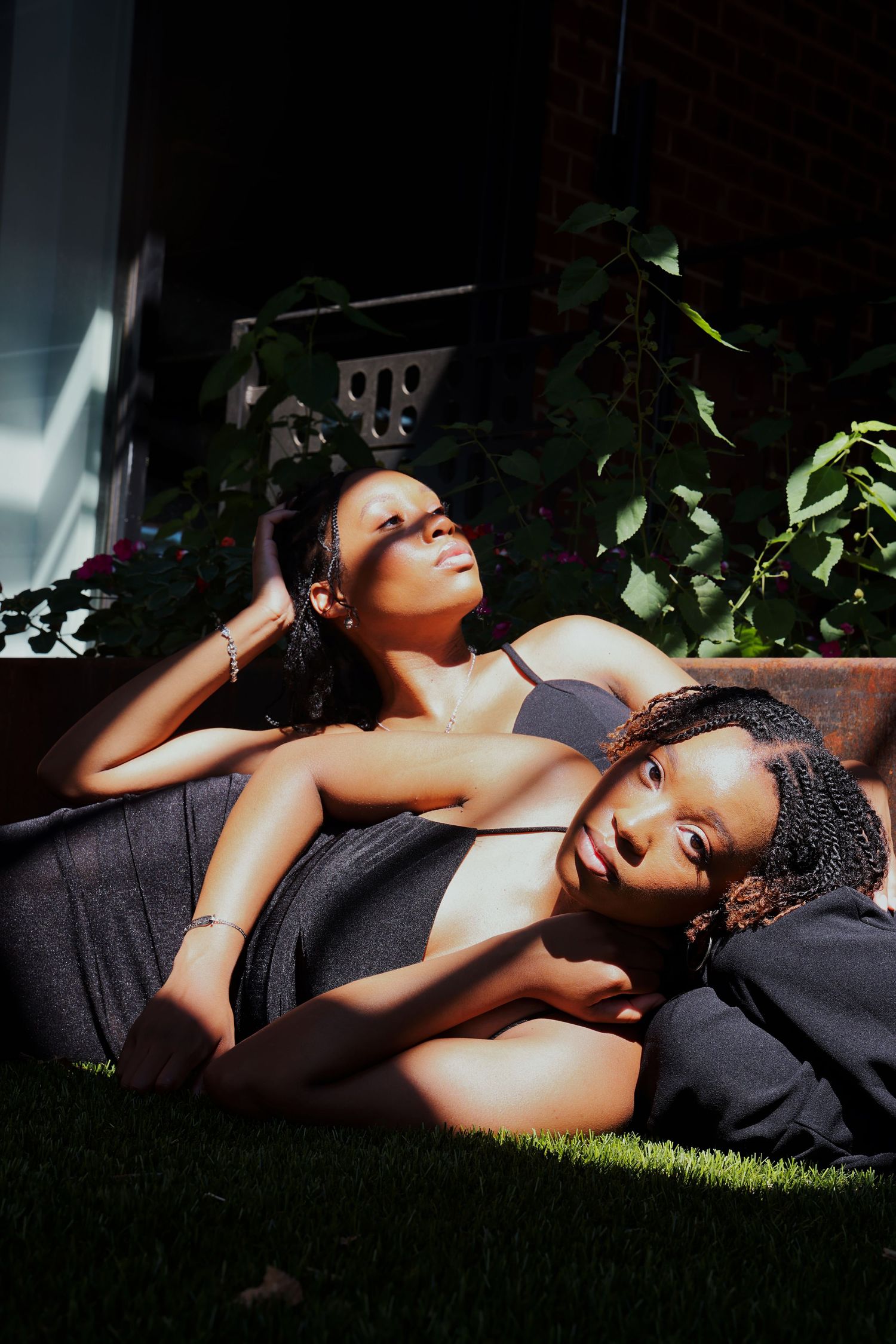 Two young women relaxing outdoors in sunlight, one resting her head on the other's lap near lush green plants.