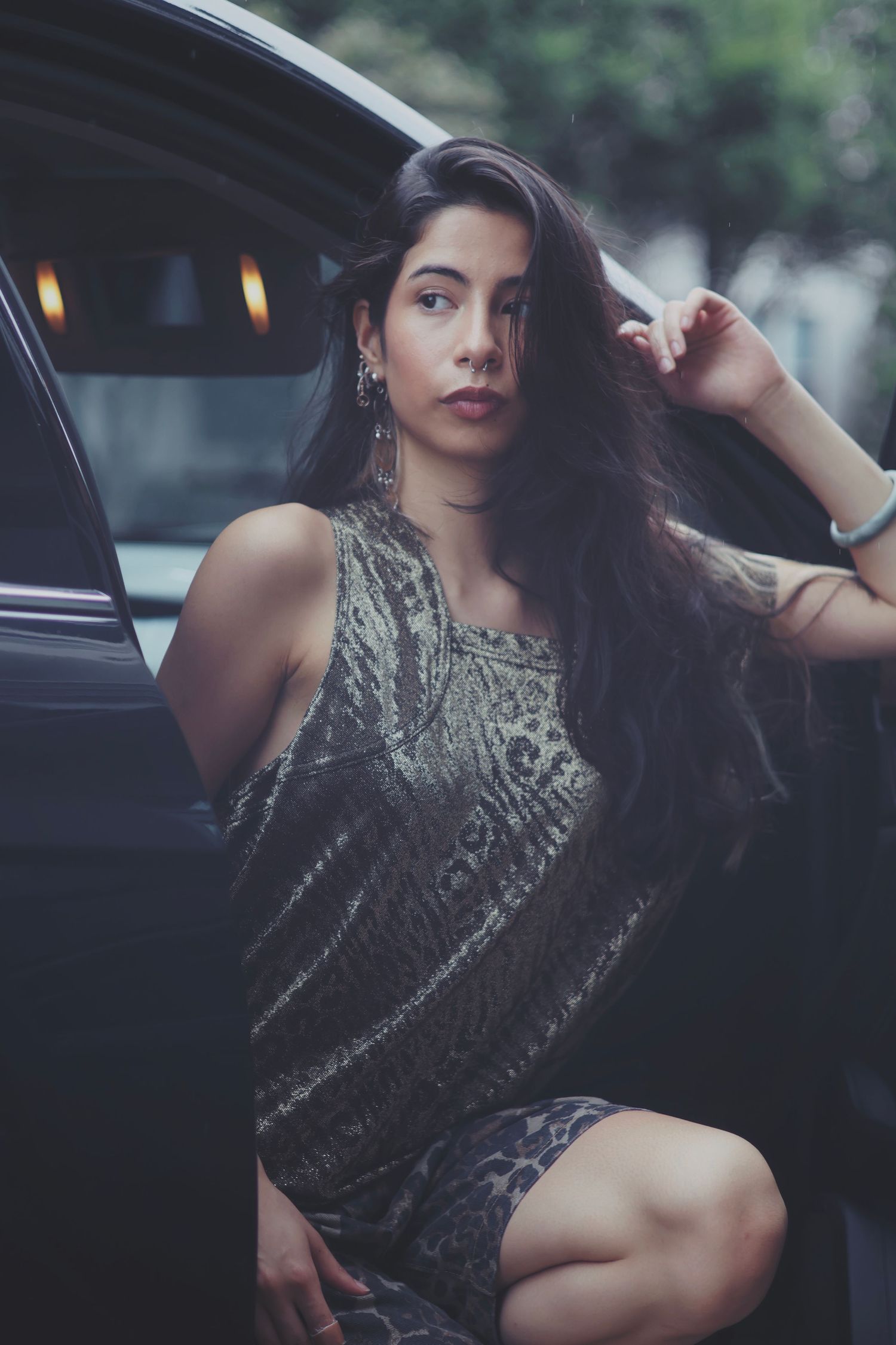 Young woman in silver dress posing by luxury car door, long dark hair, elegant street fashion photography.