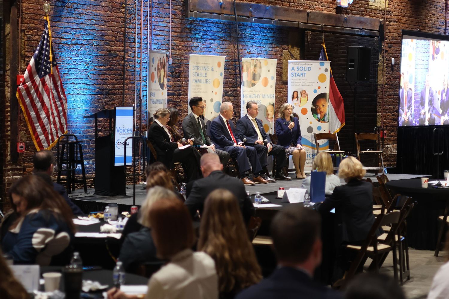 Panel discussion at a formal event in a brick venue with US flag, seated speakers facing an audience.