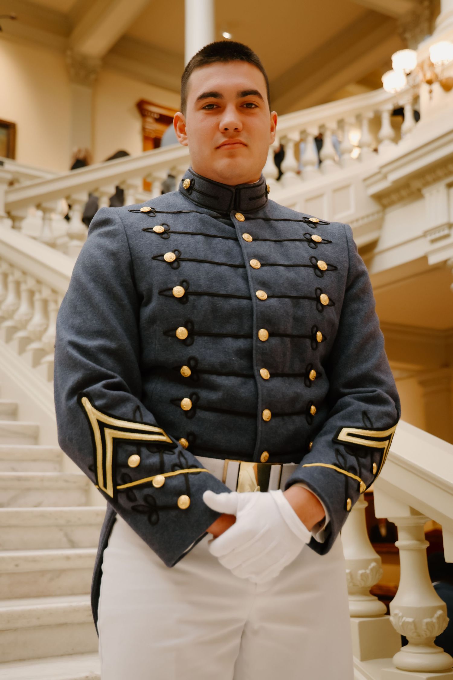 Young male cadet in formal military dress uniform with gold buttons stands on marble staircase interior.