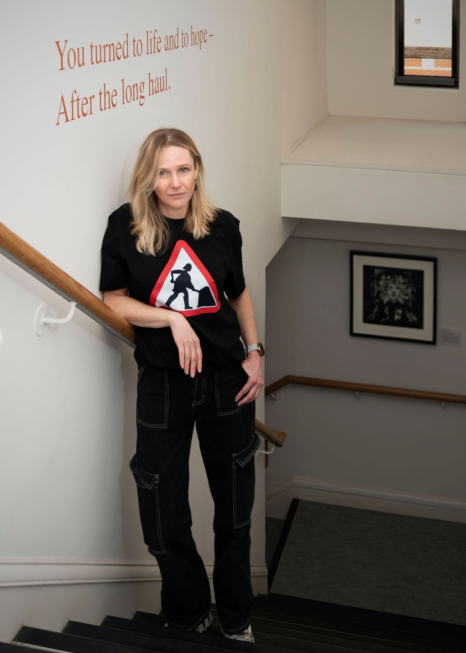 A woman, standing in a hallway under a quote from the poet Jacky Kay
