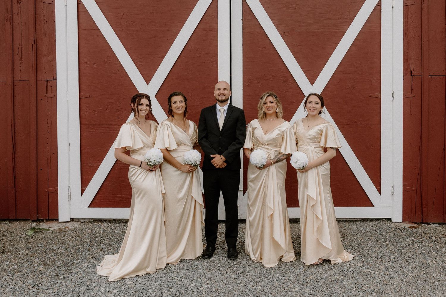 Four bridesmaids in champagne gowns and a best man in black suit pose before a red barn door.