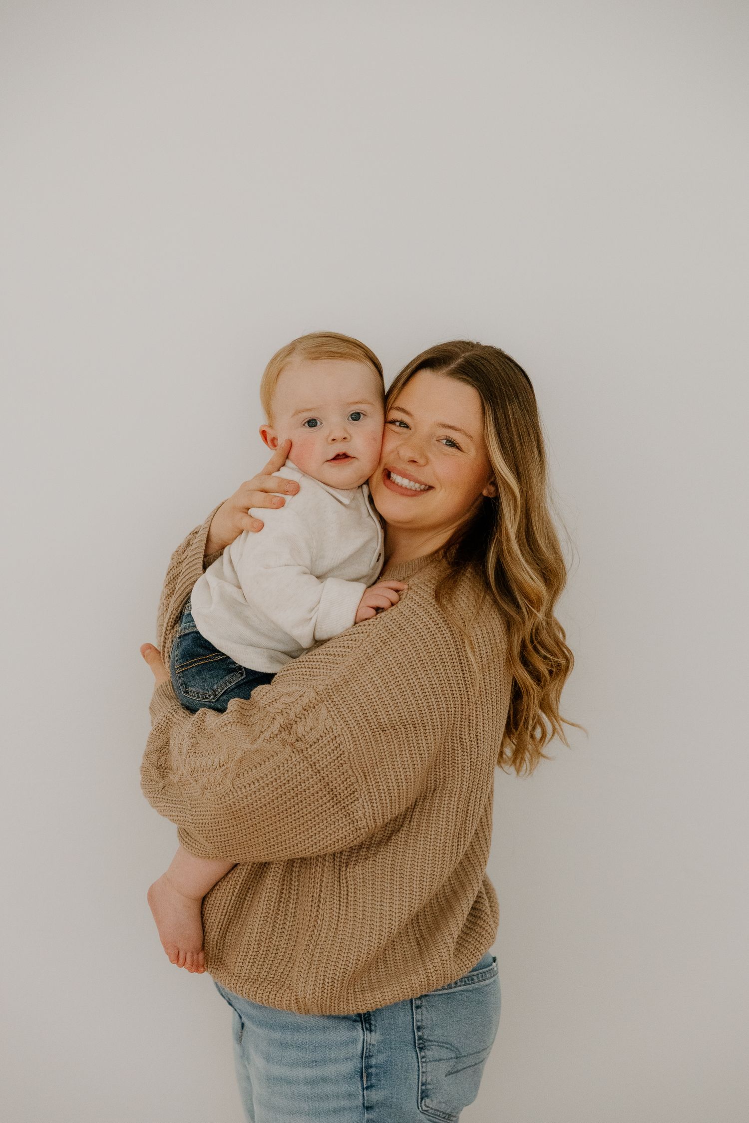 Smiling young mother in tan knit sweater holds adorable baby against a white background.