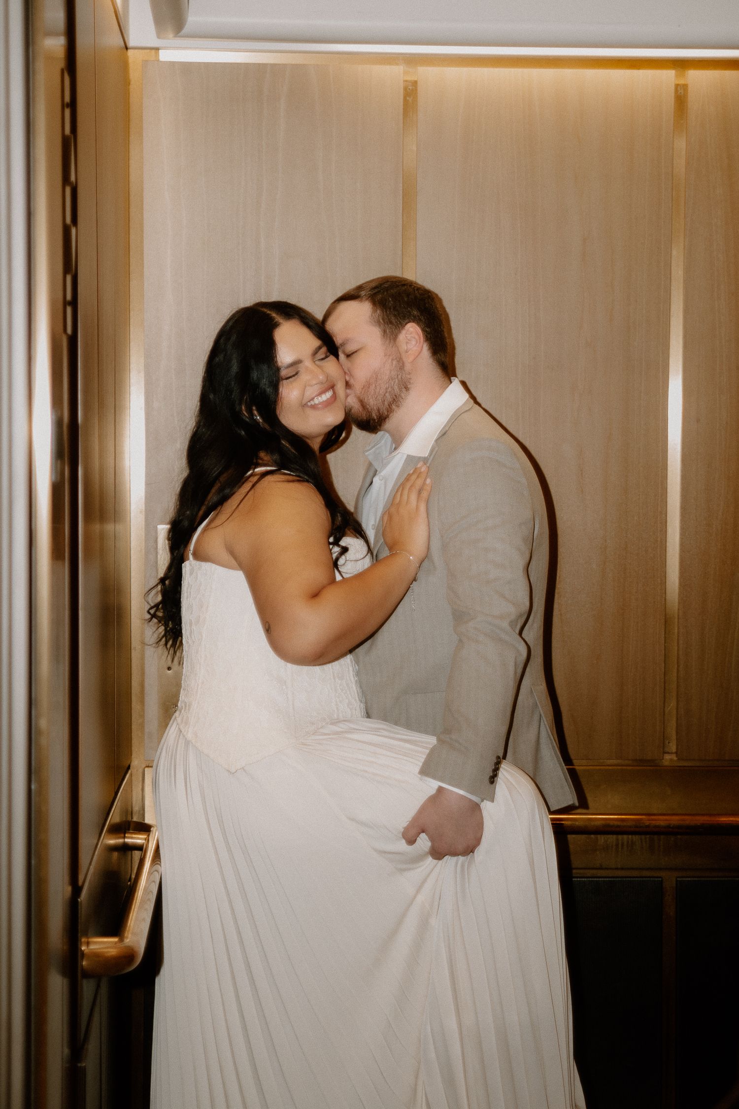 Groom kisses smiling bride on the cheek inside a warmly lit elevator, both in wedding attire.