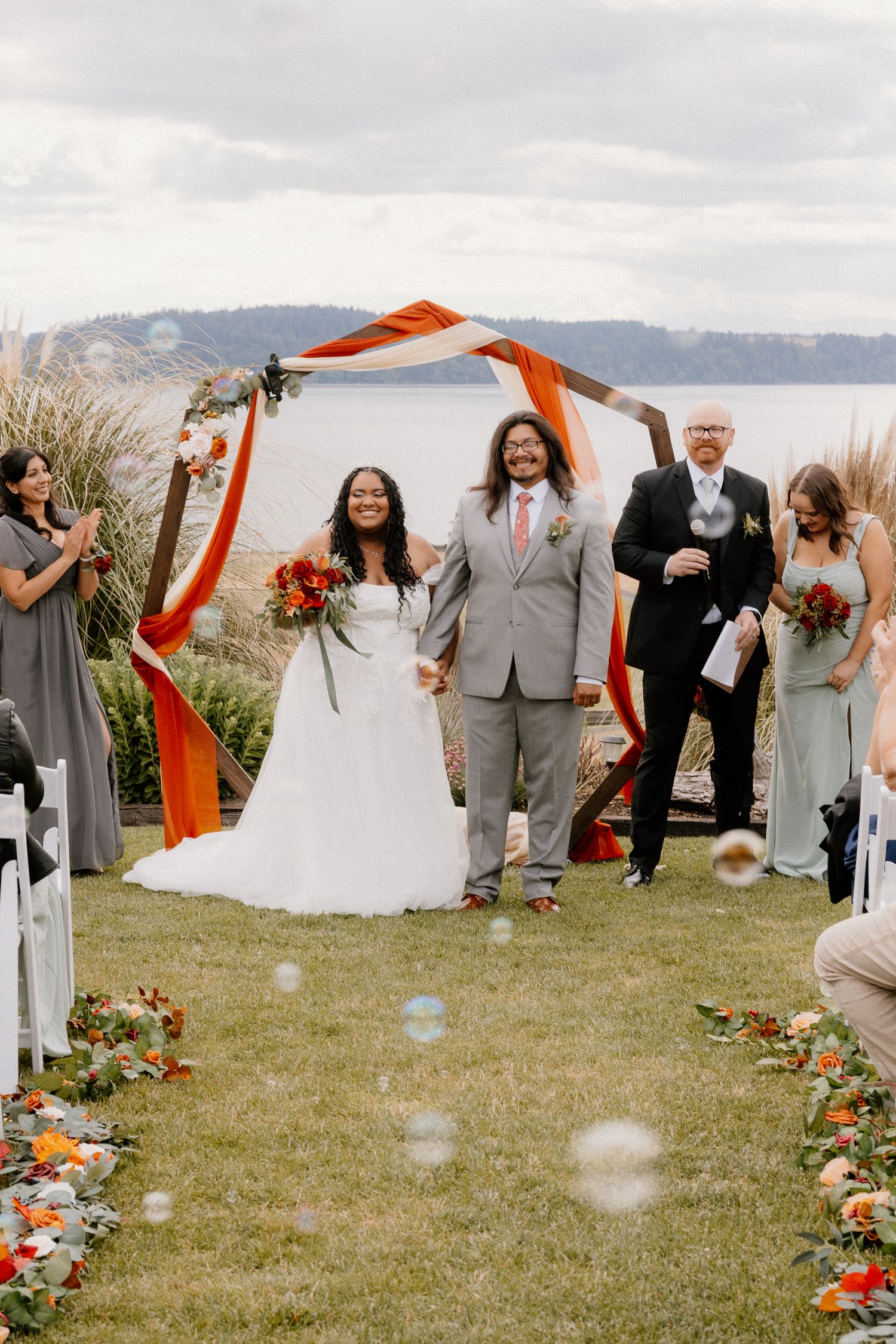 Newlyweds walk down aisle under orange draped arch with water views and bubbles floating nearby.