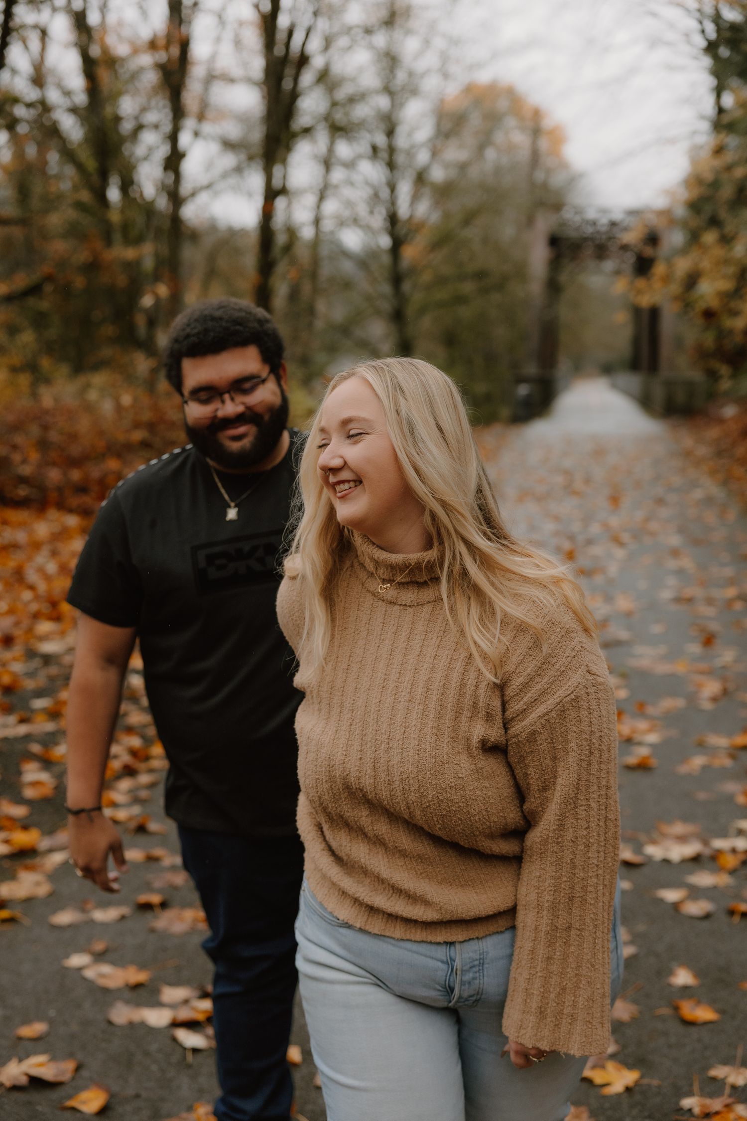 Smiling couple walking together on a leaf-covered autumn path through a wooded area.