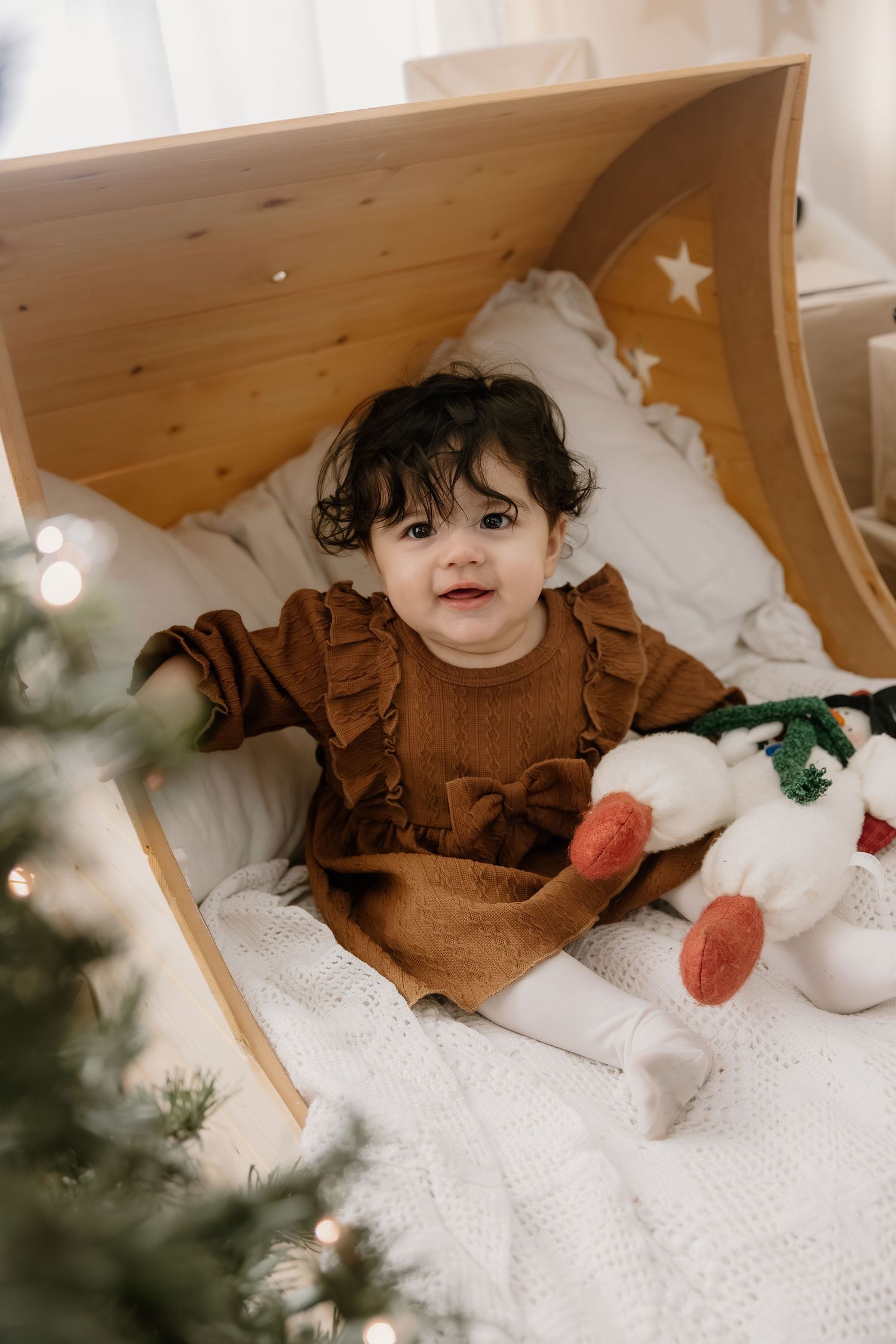 Curly-haired baby in brown dress sitting in a wooden cradle beside a Christmas tree with toys.