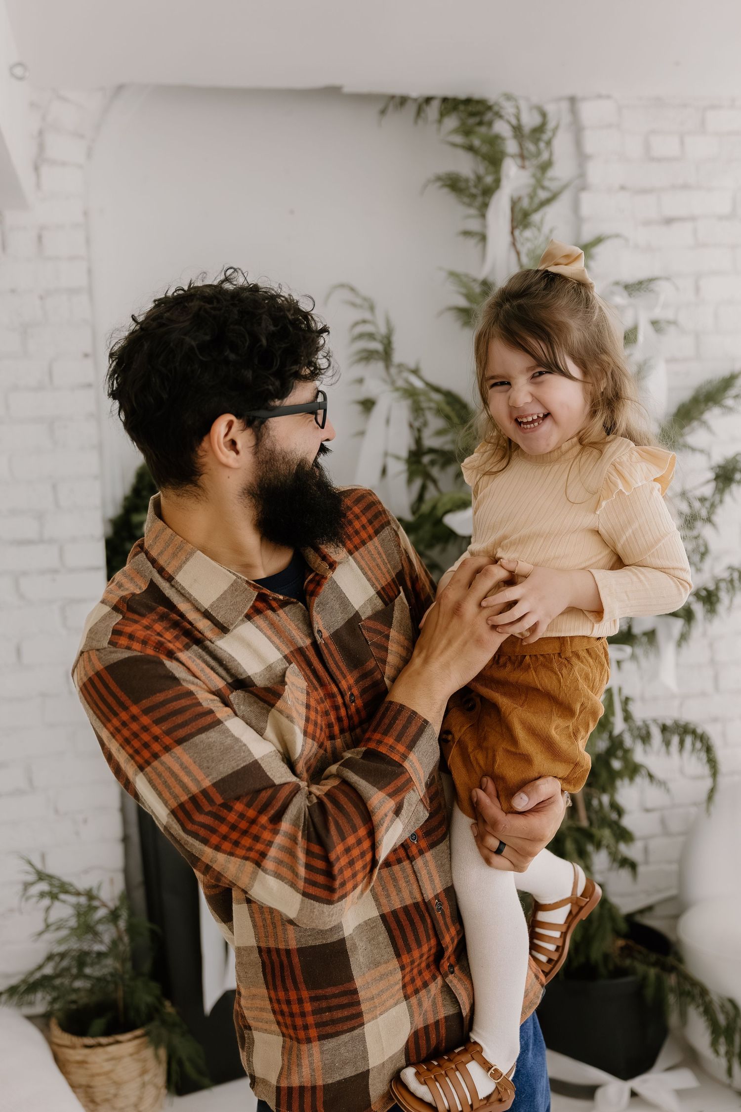 Bearded father in plaid shirt laughing while holding his giggling toddler daughter indoors.