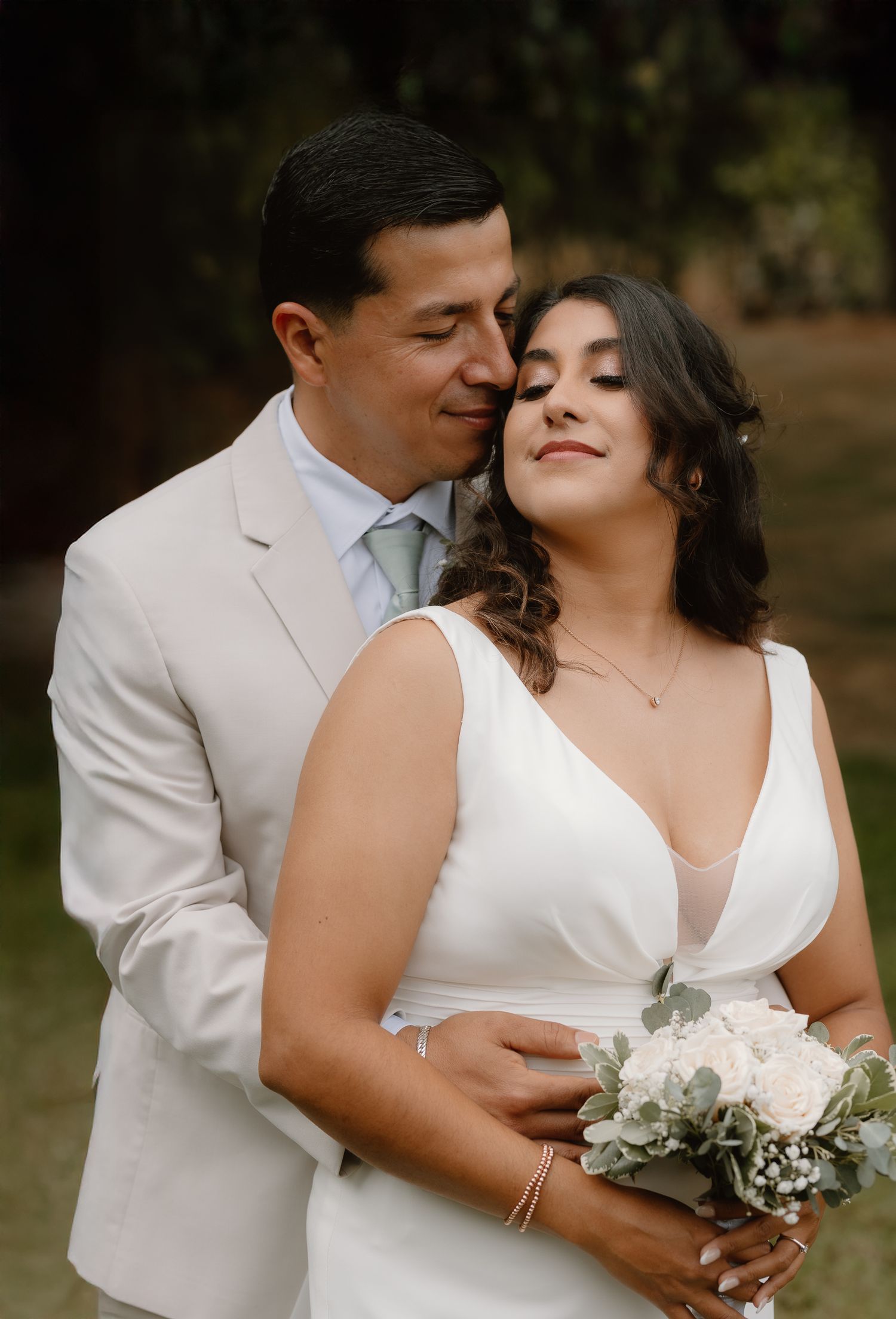 Groom in cream suit tenderly embracing bride holding white bouquet in garden setting.