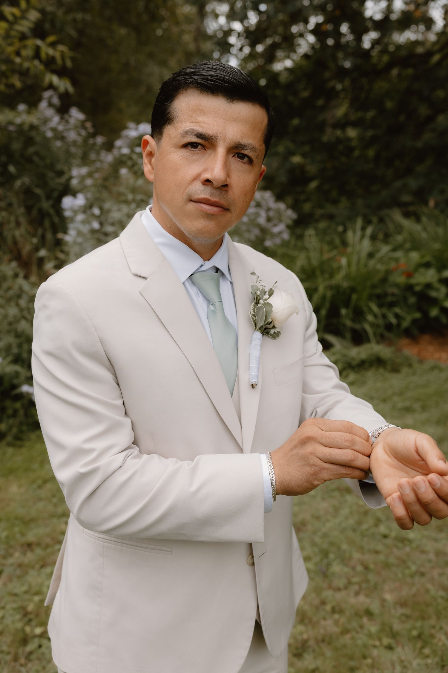 Dapper groom in beige suit adjusting cufflinks with white boutonniere in outdoor setting.