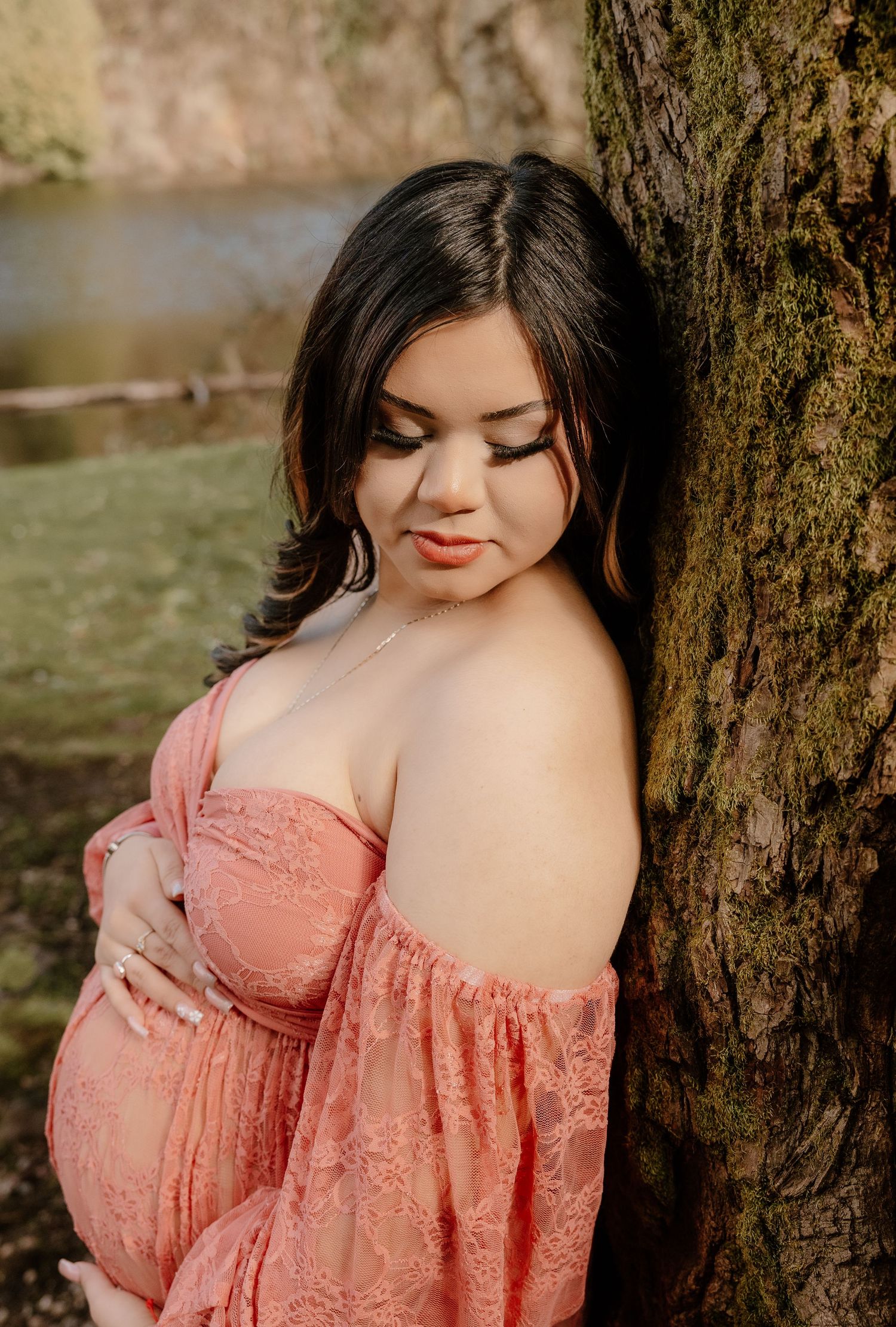 Pregnant woman in pink lace off-shoulder dress leaning against moss-covered tree with eyes closed.