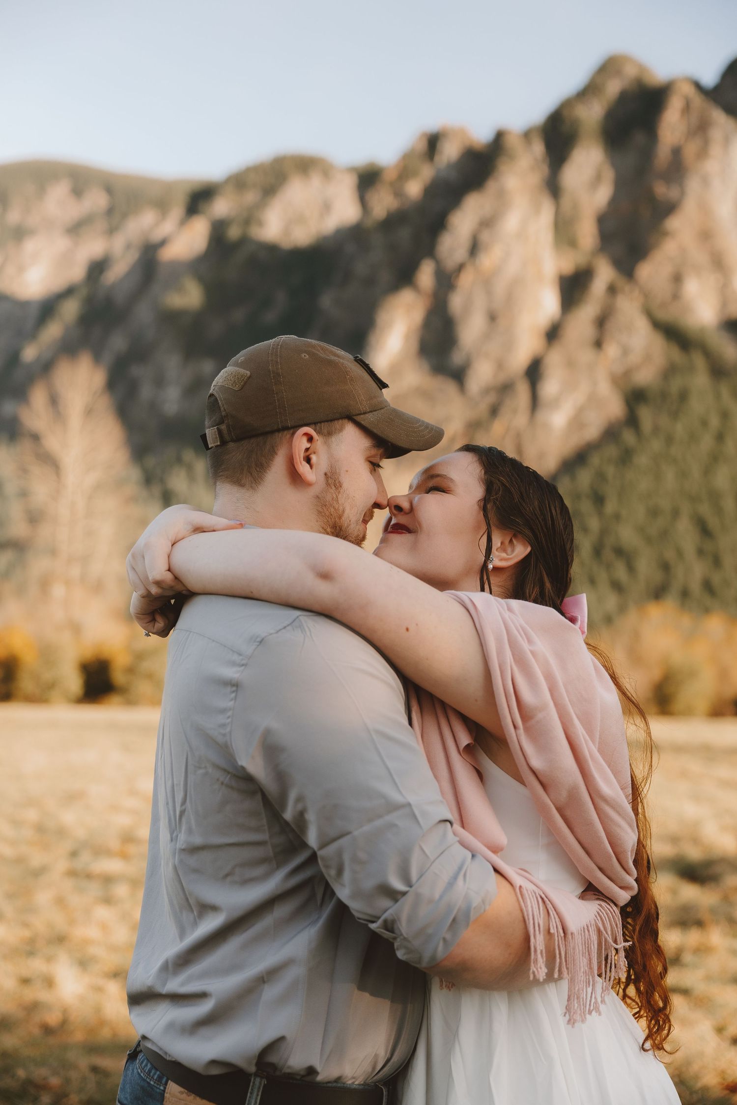 Couple embracing and almost kissing outdoors with mountain backdrop, woman in pink scarf and man in gray shirt and cap.