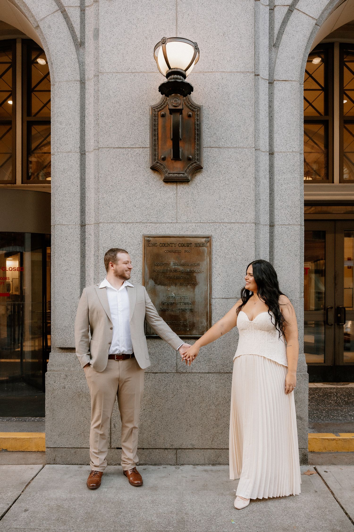 Engaged couple holds hands while standing in front of an elegant stone building with ornate lighting.