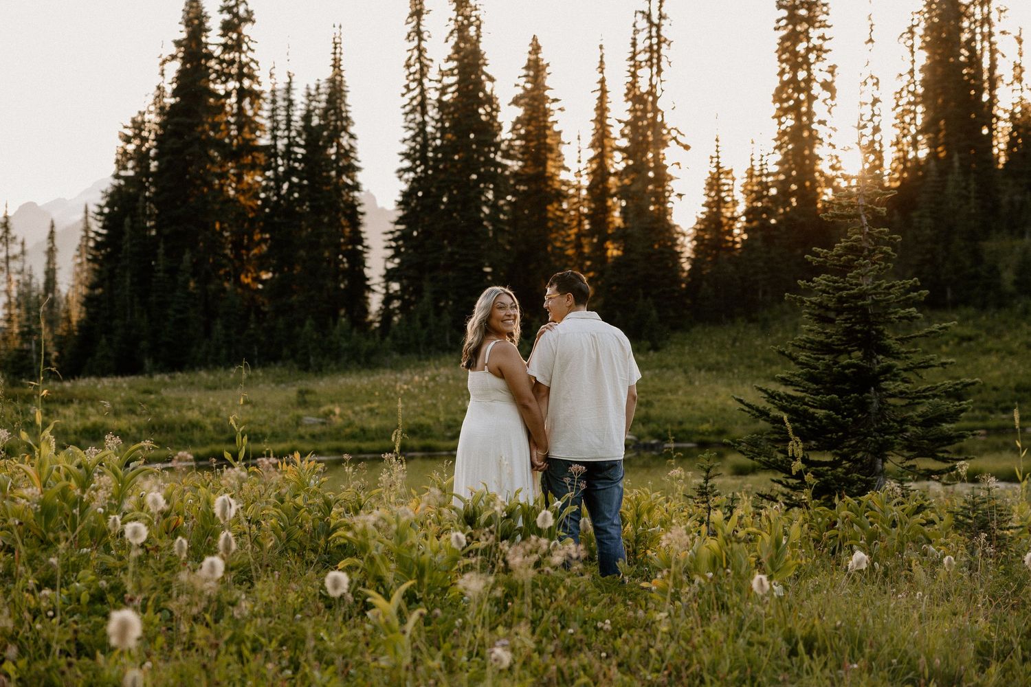 A couple holding hands in a wildflower meadow at sunset with tall evergreen trees behind them.