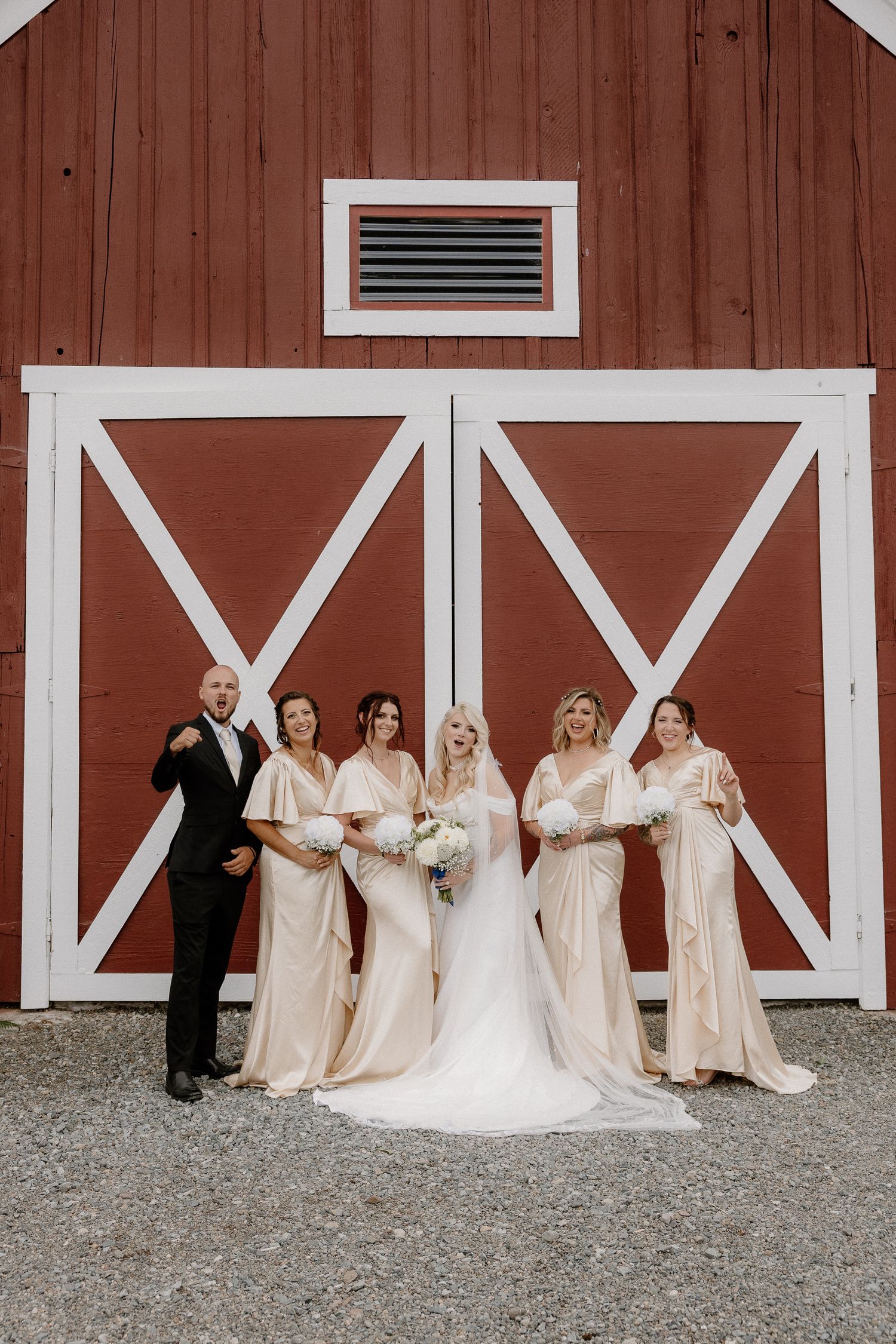 Bride with bridesmaids in champagne gowns posing in front of red barn doors at rustic wedding.