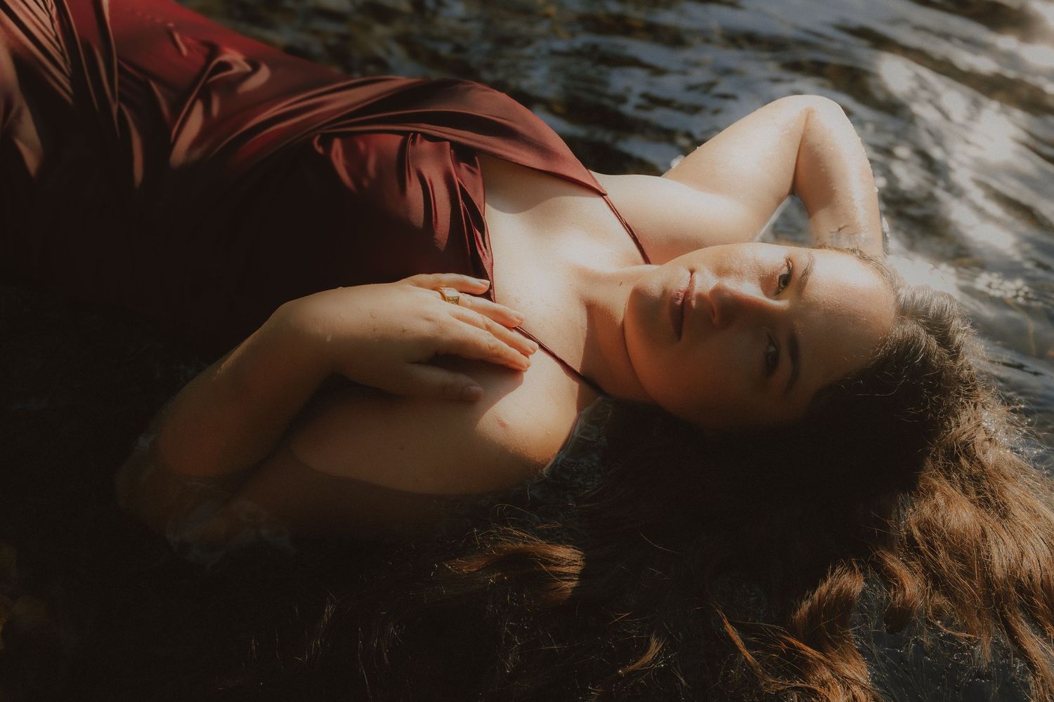 A woman in a red silk dress lies gracefully in shallow water with her hair spread around her.