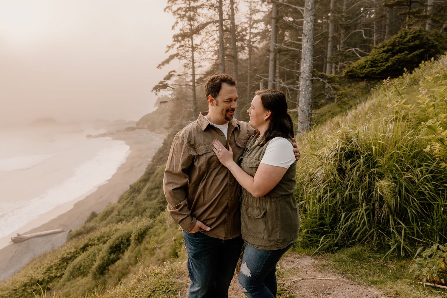 A couple stands on a coastal trail surrounded by lush greenery with a misty ocean beach backdrop.