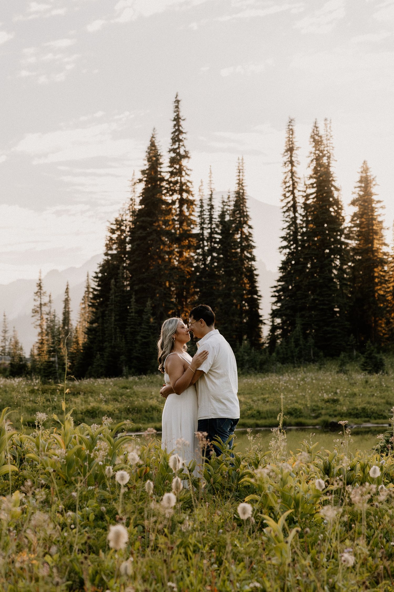 Couple embraces in a wildflower meadow at sunset with tall pine trees and mountains in the background.
