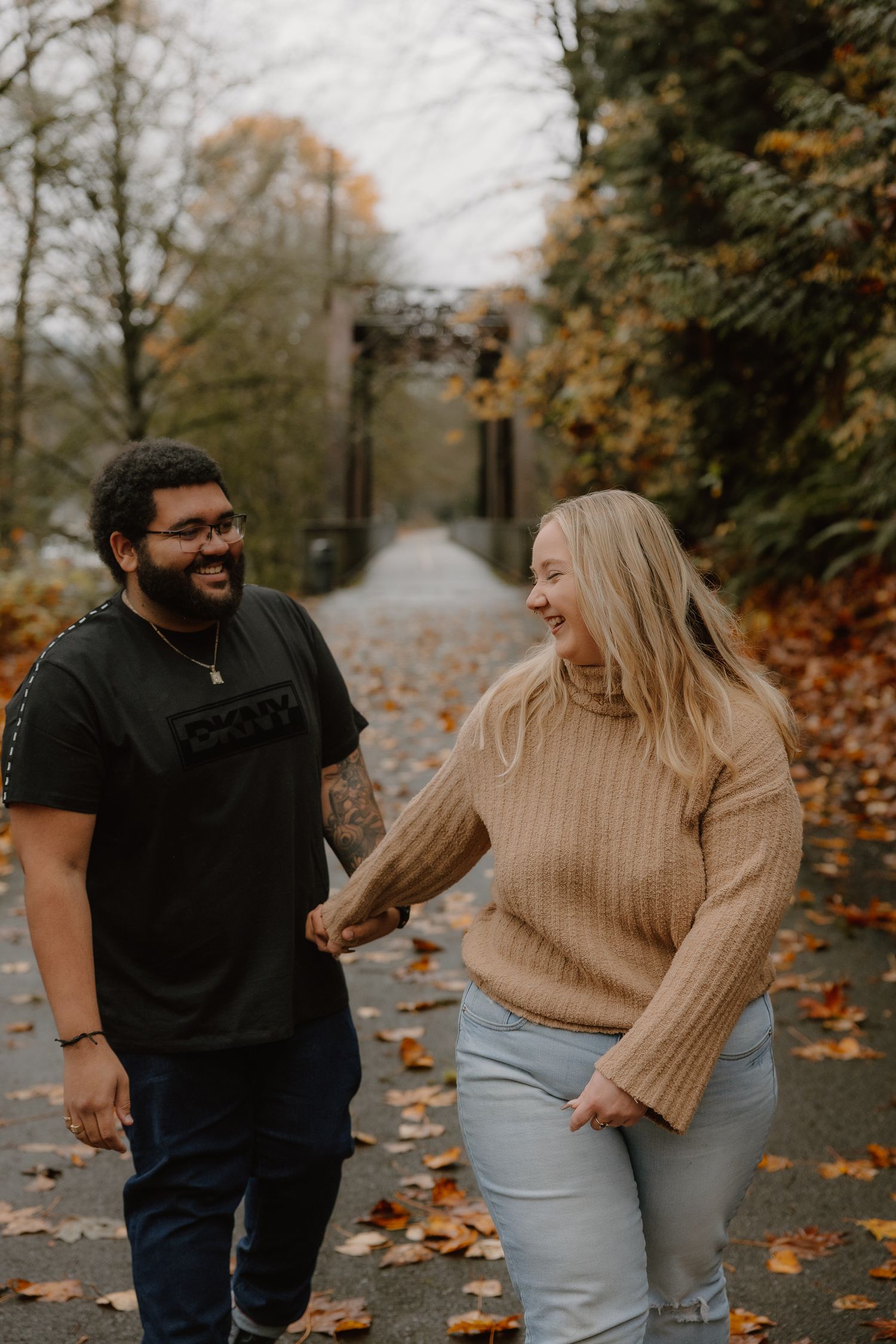 Happy couple holding hands and laughing while walking on a leaf-covered path in autumn.