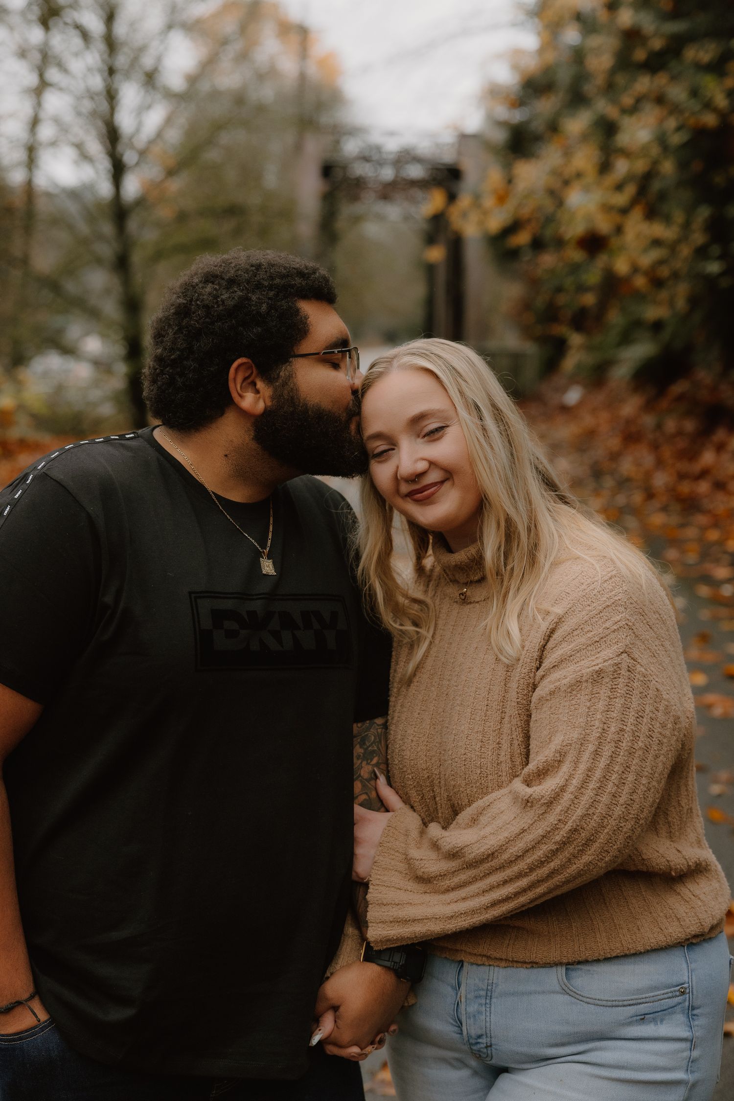 Man kissing smiling blonde woman on the cheek while holding hands on an autumn forest path.